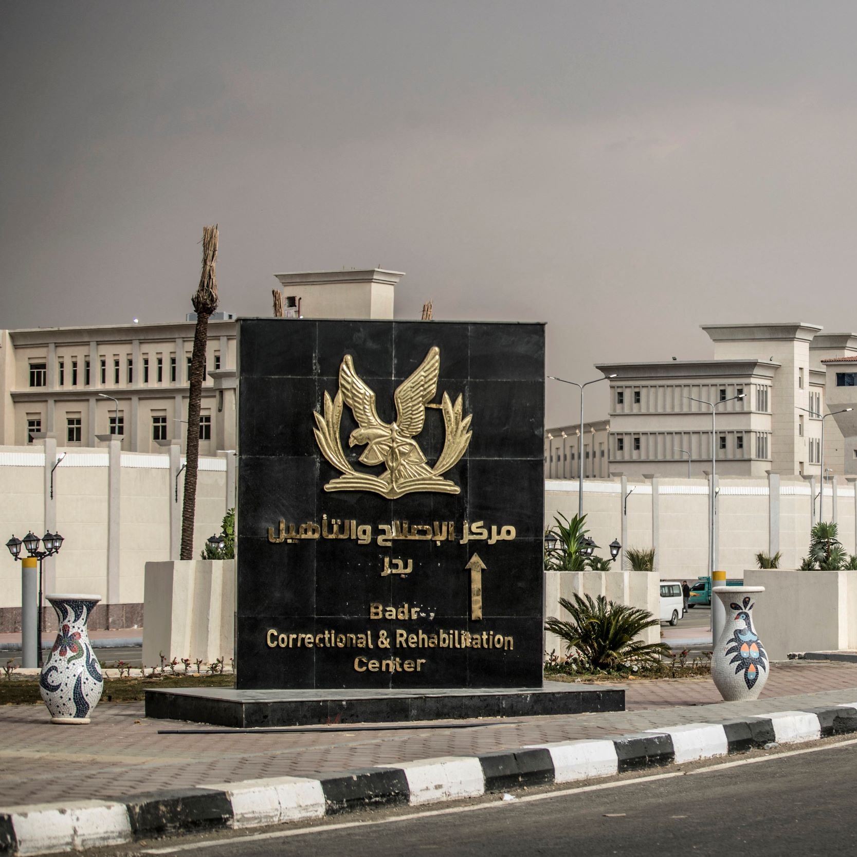 L'immagine mostra un grande cartello di benvenuto che indica il "Centro di Correzione e Riabilitazione di Badia". Il cartello è di colore scuro con un simbolo o un emblema, e si trova all'ingresso di una struttura recintata. Sullo sfondo si possono vedere edifici moderni, circondati da piante e un'illuminazione stradale. Il contesto appare ordinato e ben tenuto, suggerendo un'area istituzionale.
