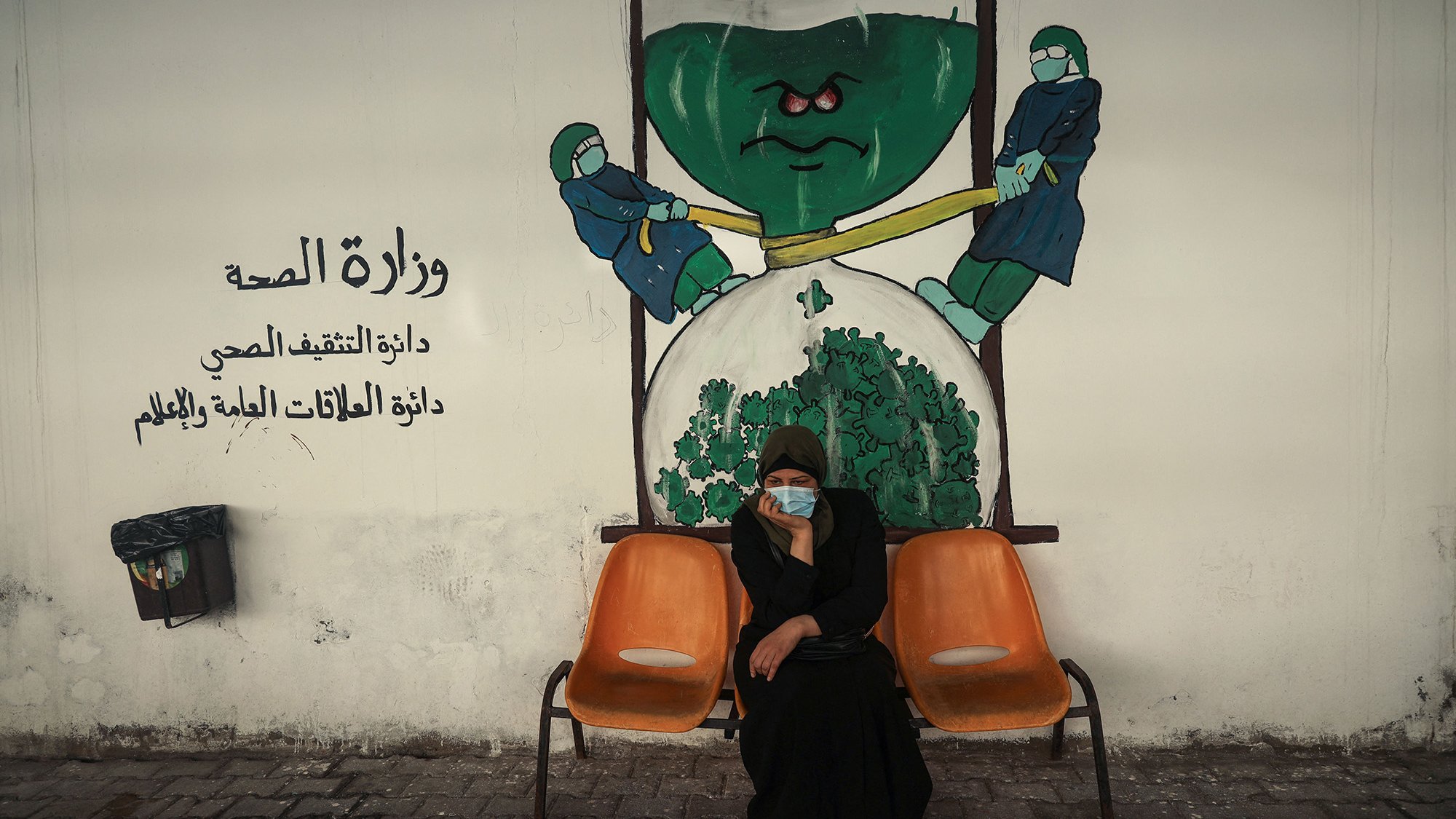 الصورة تظهر امرأة جالسة على كرسيين في منطقة تبدو وكأنها مركز صحي أو مستشفى. تحمل المرأة ملامح الجدية، وتضع كمامة على وجهها. خلفها يوجد جدار م painted يوضح رسماً يجسد فكرة وزارة الصحة، حيث يظهر شخصيتان ترتديان بزة وقاية، تسعيان لشد أو دفع شيء كبير يشبه الخضار أو الملفوف. الألوان المستخدمة في الجدار تعكس موضوع الصحة، مع تركيبات واضحة ورموز مثل القفازات والكمامات.