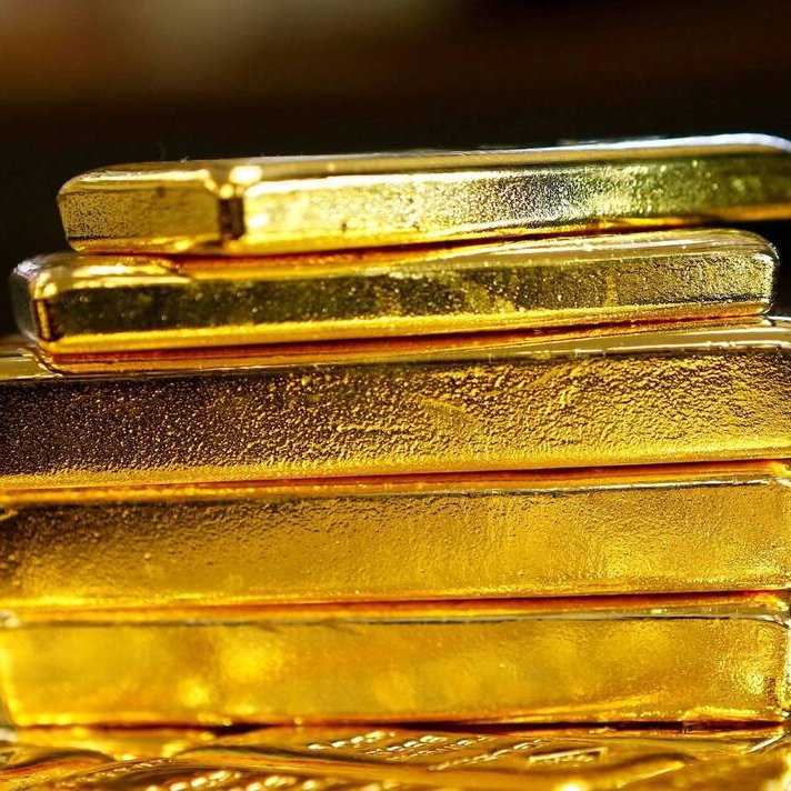 L'image montre plusieurs barres en or empilées les unes sur les autres. Chaque barre a une surface brillante et métallique, affichant la couleur dorée caractéristique de l'or. Les barres semblent bien travaillées, avec des contours nets et une texture réfléchissante. En arrière-plan, on devine une surface moins définie, ce qui met en valeur le brillant des barres d'or au premier plan.