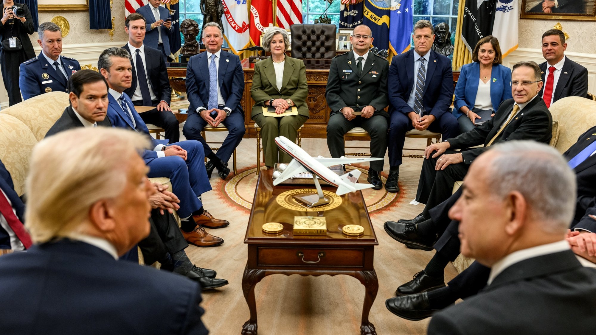 In una sala con bandiere, vari uomini e donne seduti attorno a un tavolo con un aereo in miniatura.