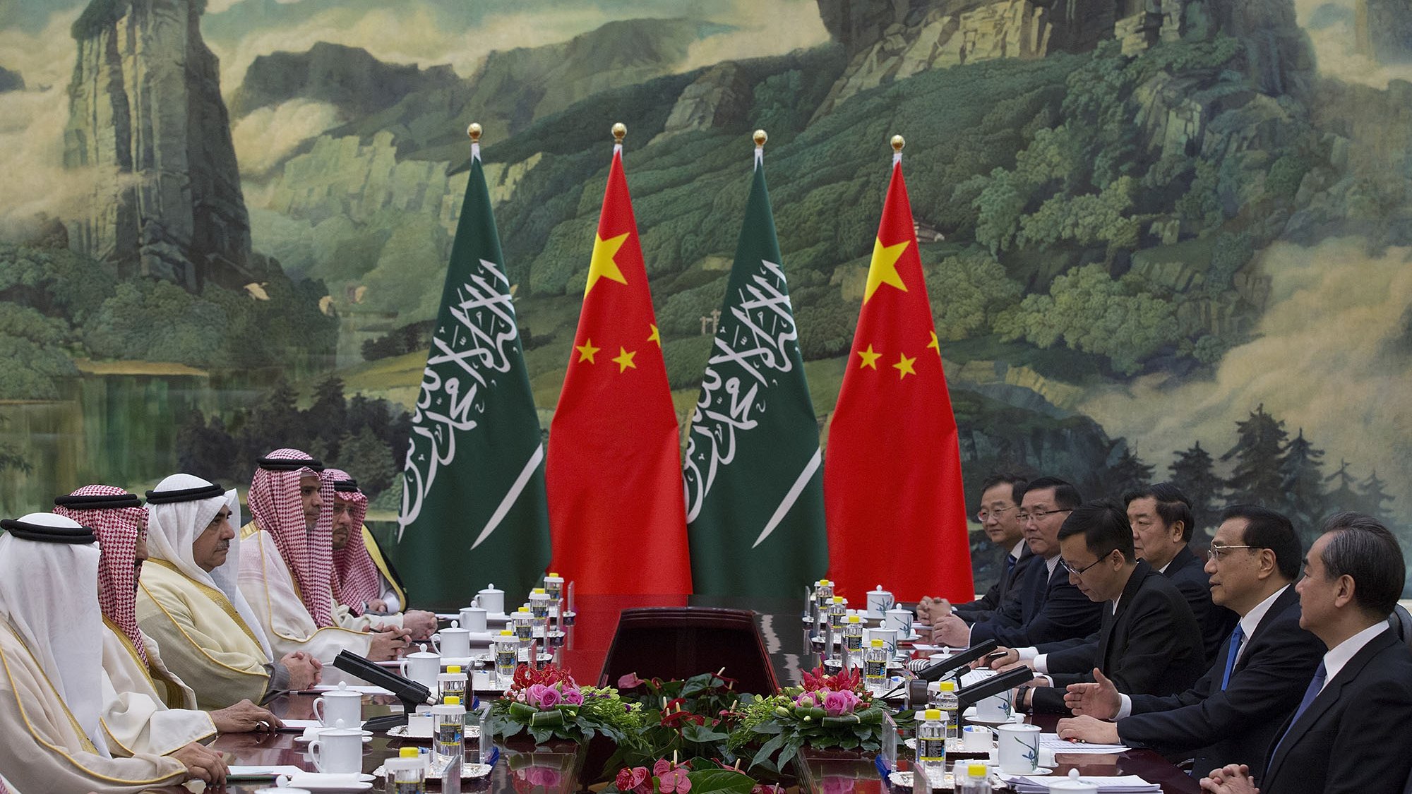 L'image montre une réunion diplomatique entre des représentants de l'Arabie saoudite et de la Chine. Les participants sont assis à une table, entourés de drapeaux des deux pays. En arrière-plan, on peut voir une grande fresque représentant des paysages naturels. La scène dégage une ambiance formelle, typique des sommets ou des négociations internationales. Sur la table, il y a des documents et des tasses de thé.