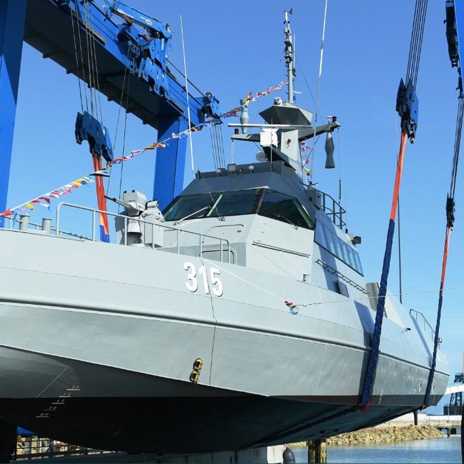 L'image montre un navire militaire en cours de manutention. Il est positionné sur un quai, soutenu par des grues bleues qui le soulèvent avec des sangles. Le bateau est de couleur grise et porte le numéro 315. Le ciel est clair, suggérant une journée ensoleillée. Cette scène évoque des opérations de maintenance ou de construction navale.