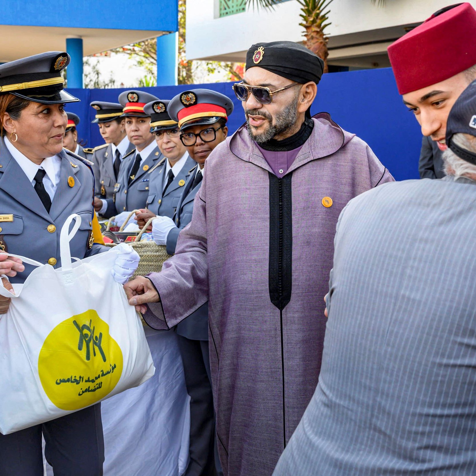 الصورة تظهر مجموعة من الأشخاص في مناسبة رسمية. يبدو أن هناك توزيعًا للأغراض، حيث تظهر سيدة ترتدي uniform رسمي وتحمل حقيبة. الشخص في الوسط يرتدي جلبابًا تقليديًا ونظارات شمسية، ويبدو أنه يقوم بمصافحة أحد الأشخاص. في الخلفية، يلاحظ وجود رجال ونساء يرتدون زيًا رسميًا، مما يوحي بأن الحدث له طابع رسمي أو احتفالي. الأجواء تبدو مفعمة بالنشاط والترحيب.