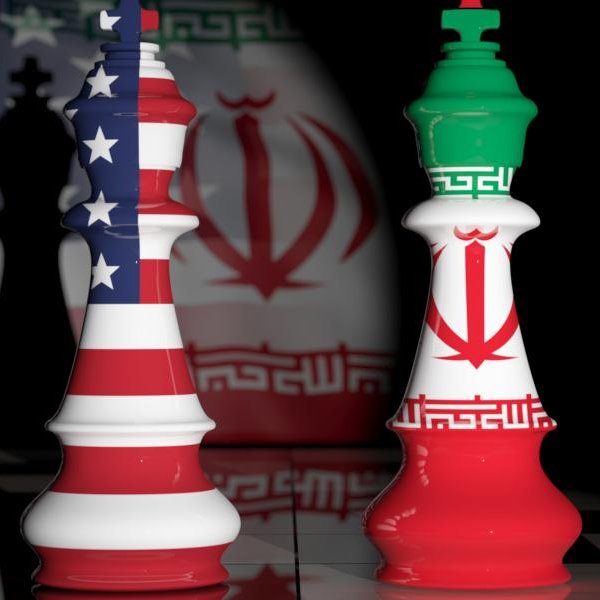 L'image représente des pièces d'échecs en forme de rois, une avec les couleurs du drapeau américain et l'autre avec celles du drapeau iranien. En arrière-plan, on peut voir des ombres ou des motifs représentant ces deux pays. La scène évoque une confrontation symbolique entre les États-Unis et l'Iran, souvent associée à des tensions géopolitiques. Le fond sombre renforce l'aspect dramatique de l'image.