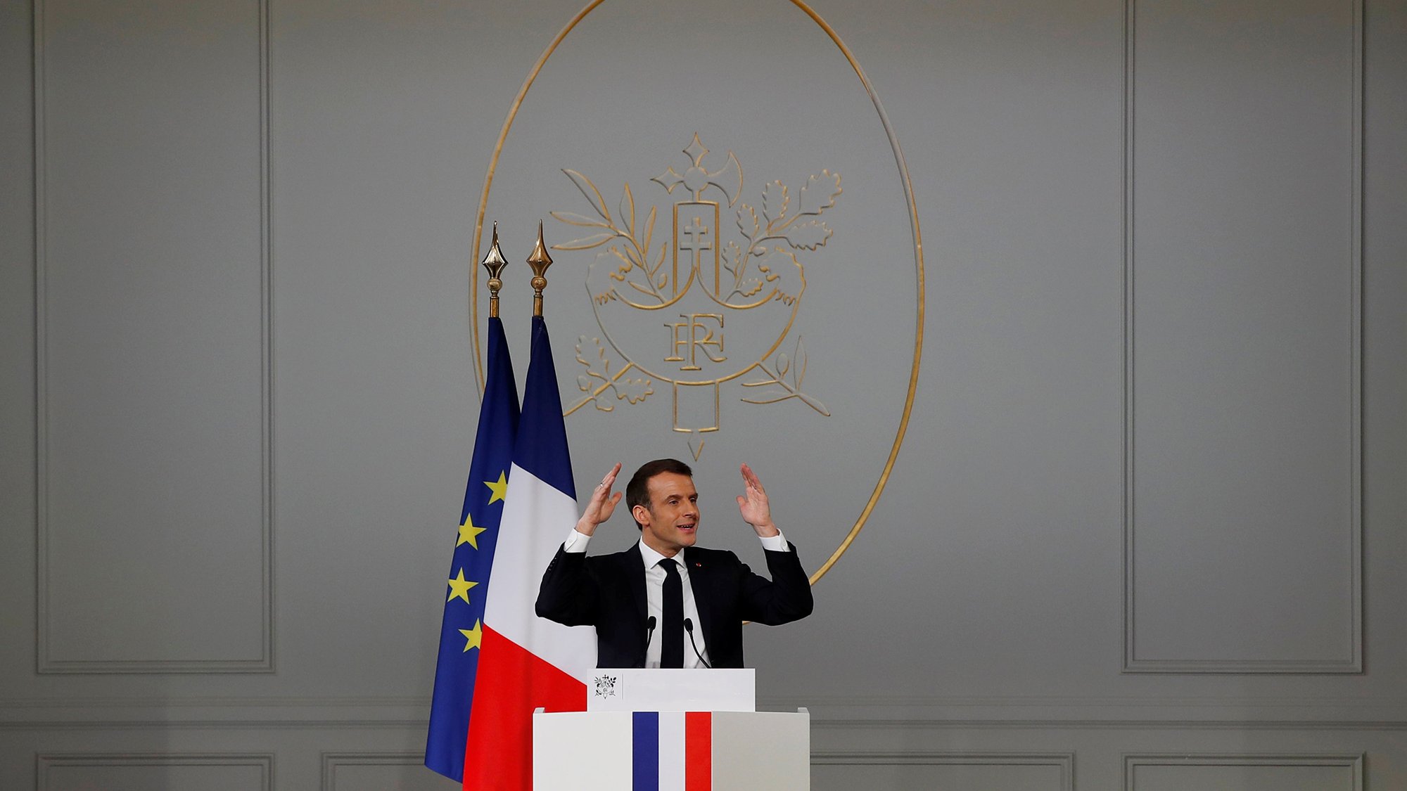 L'image montre un homme debout derrière un pupitre, avec les mains levées comme s'il s'adressait à un auditoire. Derrière lui, il y a un drapeau français et un symbole décoratif sur le mur. Le pupitre présente des couleurs bleu, blanc et rouge, rappelant celles du drapeau français. L'intérieur semble officiel, avec des murs clairs et une ambiance formelle. L'homme semble être en train de faire un discours ou une déclaration importante.