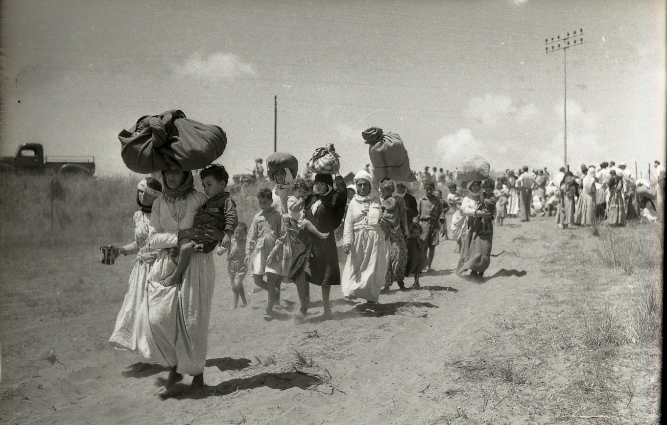 L'image montre un groupe de personnes marchant le long d'un chemin poussiéreux. Certaines femmes portent de lourdes charges sur la tête, tandis que d'autres tiennent des enfants par la main. La scène semble illustrer un moment de déplacement ou de migration, avec des paysages arides en arrière-plan. Des nuages blancs ponctuent le ciel, et on peut voir des lignes électriques au loin. L'atmosphère de la photo évoque la lutte et la détermination des personnes en marche.