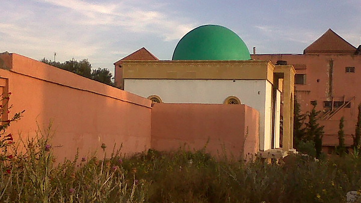 L'image montre un bâtiment avec une coupole verte, partiellement cachée par un mur. Le bâtiment semble être de style architectural traditionnel, avec des murs peints en rose et des fenêtres rondes. On remarque également une végétation sauvage devant, ajoutant un aspect naturel à la scène. En arrière-plan, on aperçoit d'autres structures, suggérant un environnement urbain ou semi-urbain. Le ciel est clair, indiquant une atmosphère paisible.