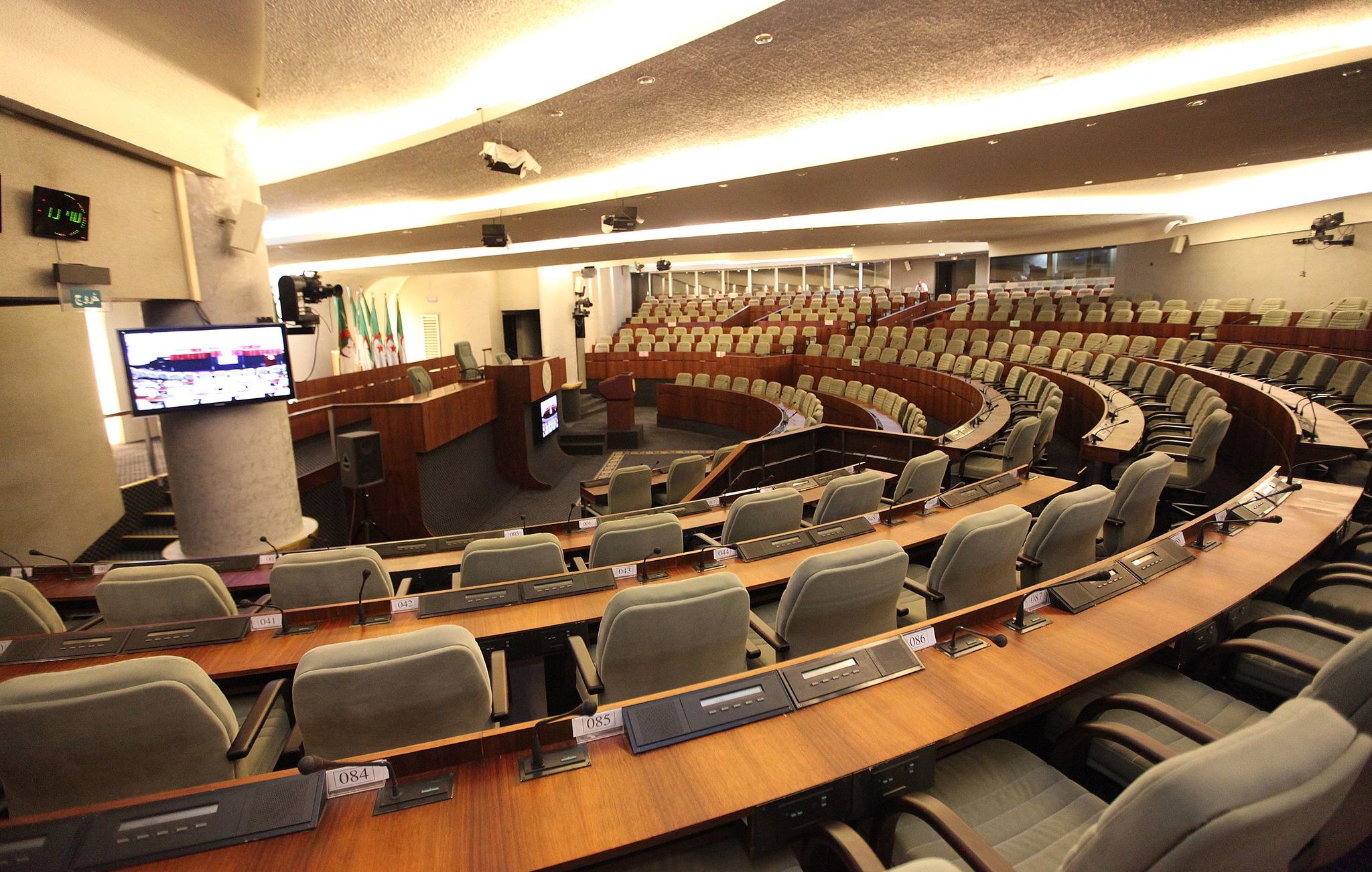 L'image montre une salle de réunion ou de débat, probablement une chambre législative. On y voit plusieurs rangées de sièges grisés disposés en arc de cercle autour d'une scène centrale. À l'avant, un bureau ou une tribune est visible, avec des équipements audiovisuels, comme un écran et des micros intégrés dans les tables. L'éclairage est doux et moderne, créant une atmosphère propice aux discussions. Les murs sont revêtus de matériaux contemporains, donnant une impression de sérieux et de fonctionnalité.