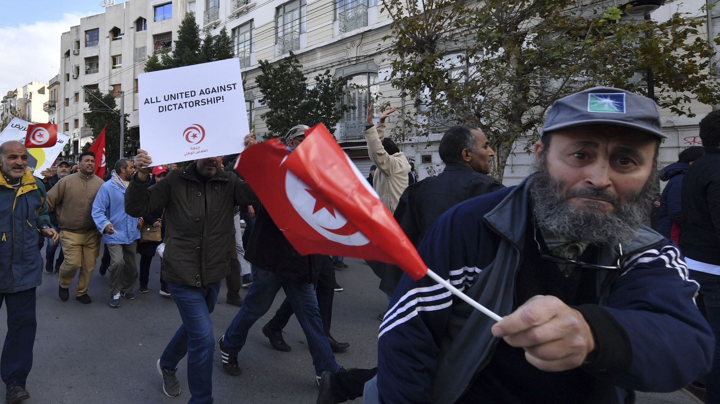 L'image montre une scène de manifestation avec des gens défilant dans la rue. Plusieurs manifestants portent des drapeaux tunisiens et tiennent des panneaux, dont l'un exprime un message contre la dictature. L'atmosphère semble énergique et engagée, avec des participants qui expriment leurs opinions et cherchent à faire entendre leur voix. Les bâtiments en arrière-plan reflètent un contexte urbain.