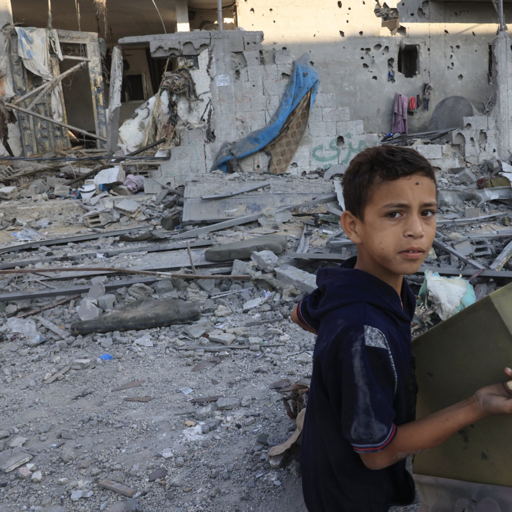 این تصویر یک پسر جوان را نشان می‌دهد که در میان ویرانه‌های یک ساختمان آسیب‌دیده ایستاده است. او در حال حمل یک جعبه است و به سمت دوربین نگاه می‌کند. محیط اطراف او پر از زباله، آوار و تکه‌های مصالح ساختمانی است. دیوارهای خراب و نشانه‌های تخریب در پس‌زمینه مشهود است و وضعیت کلی تصویر نشان‌دهنده یک منطقه آسیب‌دیده از جنگ یا بلایای طبیعی است. احساساتی نظیر ناامیدی و تلخی در چهره کودک منعکس شده است.