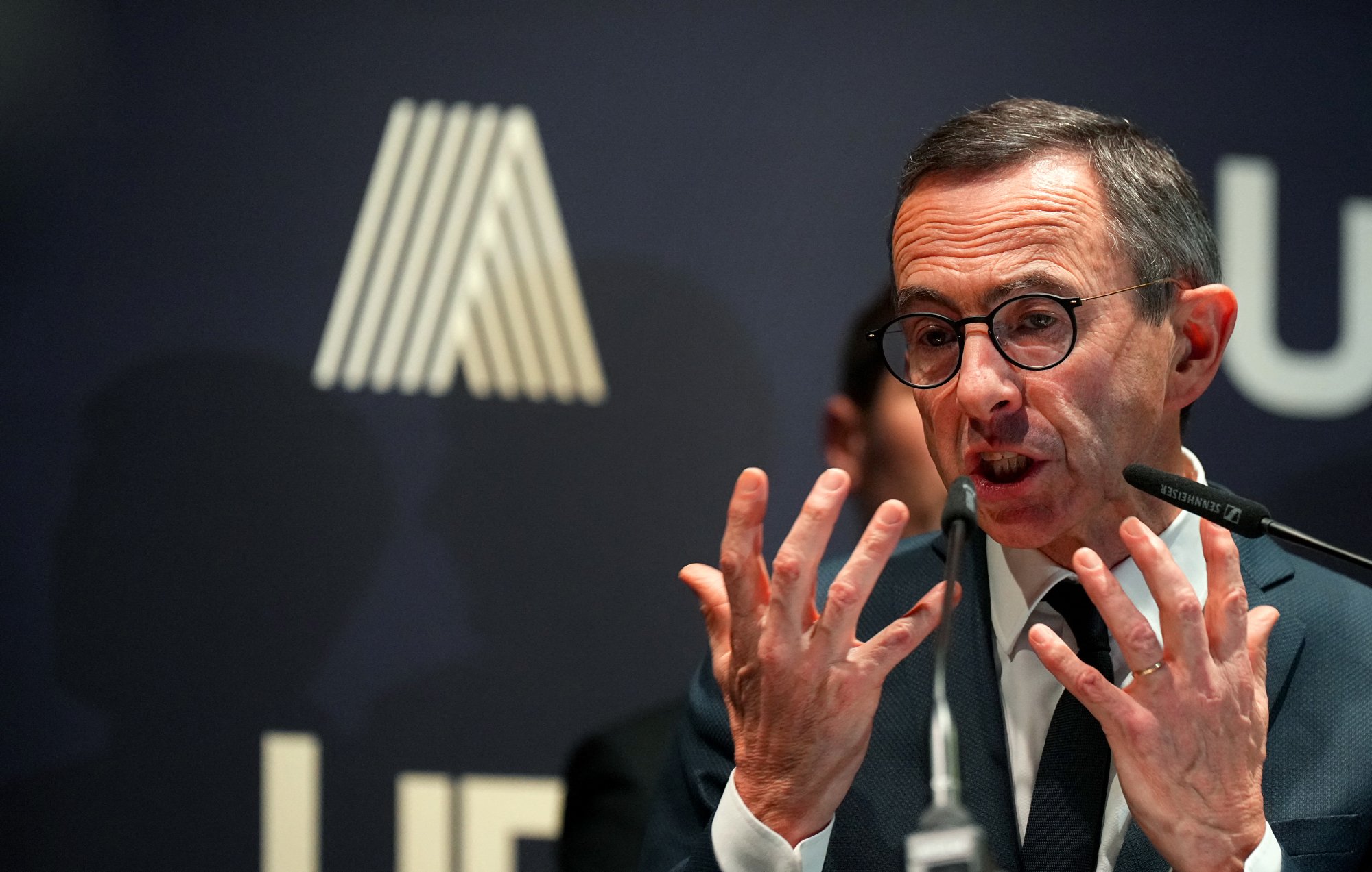 الصورة تظهر شخصًا يرتدي نظارات ويبدو أنه يتحدث بحماس، حيث يستخدم يديه للتعبير عن أفكاره. خلفه توجد خلفية داكنة تحمل شعار يبدو أنه مرتبط بمؤسسة أو منظمة. الأضواء تركز عليه مما يعطي انطباعًا بأهمية الحدث الذي يتحدث فيه.