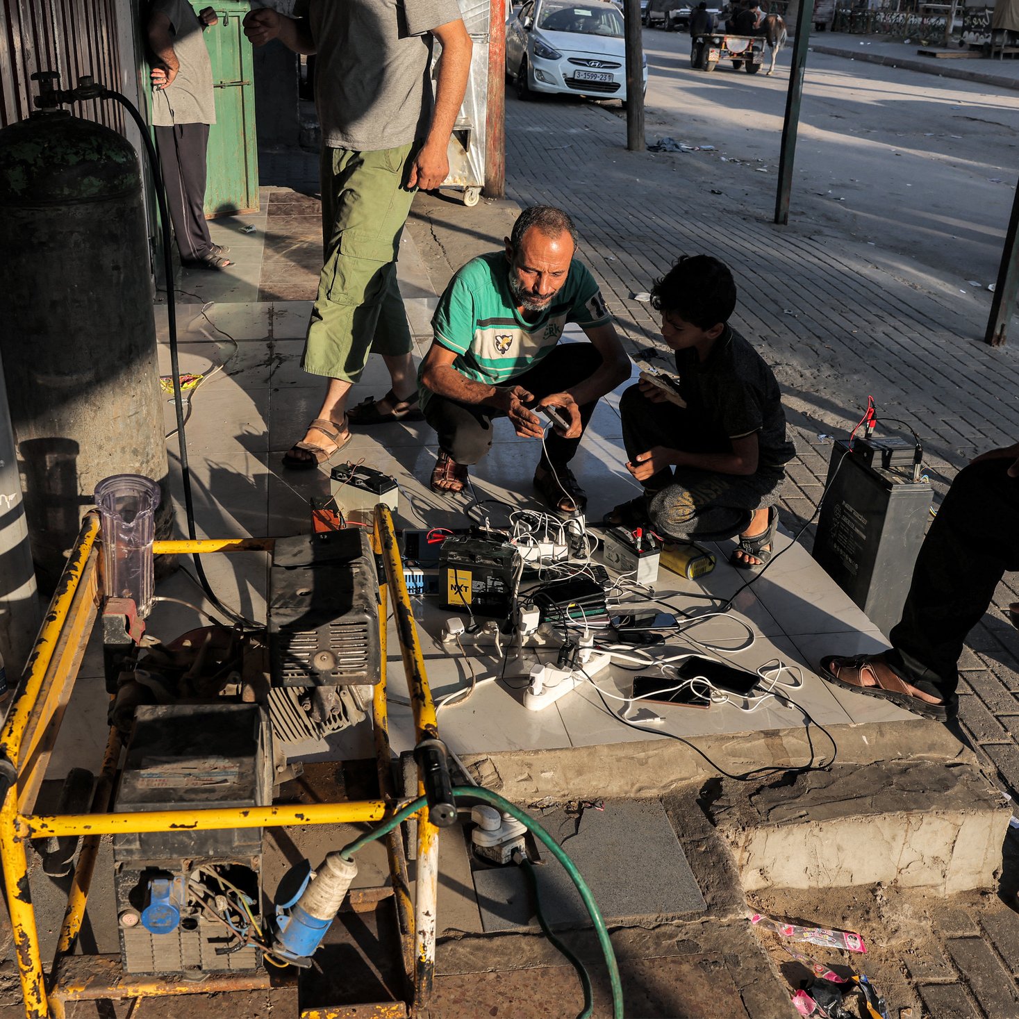 Cette image montre un groupe de personnes, principalement des hommes, rassemblées autour d'un générateur et de plusieurs chargeurs de téléphone portable. Ils se trouvent à l'extérieur, probablement sur un trottoir ou une zone piétonne pavée. Plusieurs téléphones portables sont connectés aux chargeurs qui sont branchés au générateur. À l'arrière-plan, on peut voir une voiture garée et des bâtiments. L'atmosphère semble indiquer une situation où les gens utilisent un générateur pour charger leurs appareils en l'absence d'une source d'électricité régulière.