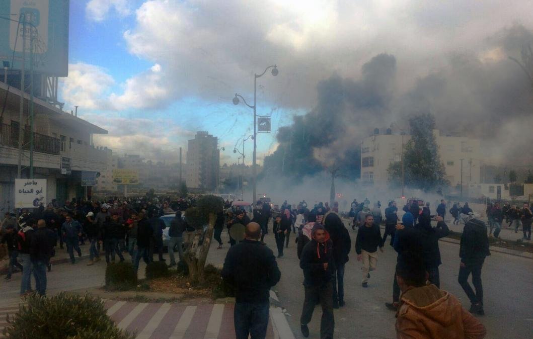 L'image montre une scène de manifestation dans une ville, avec une foule rassemblée dans une rue. Il y a des nuages de fumée et des signes de tension, suggérant des troubles ou des affrontements. Les gens marchent, certains semblent fuir la fumée, tandis que des bâtiments et des arbres sont visibles en arrière-plan. Le ciel est partiellement nuageux, ajoutant à l'atmosphère dramatique de la scène.