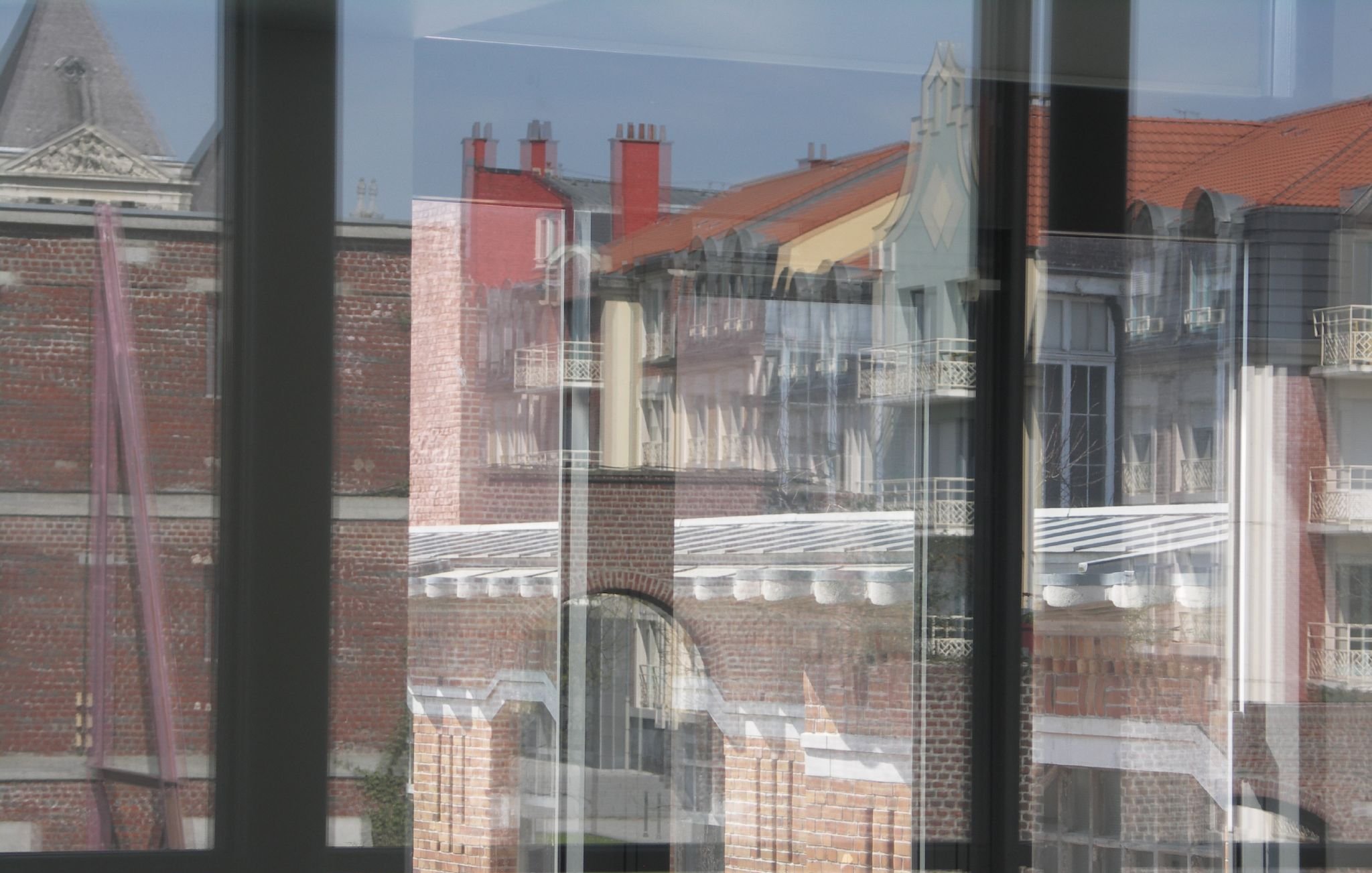 این تصویر نمایی از ساختمان‌ها را از طریق شیشه‌های یک ساختمان دیگر نشان می‌دهد. شیشه‌ها باعث ایجاد انعکاس‌هایی از بیرون می‌شوند که موجب می‌شود جزئیات معماری و رنگ‌های دیوارها و بام‌ها به خوبی نمایان شود. در پس‌زمینه، سقف‌های شیب‌دار و ساختمان‌های مختلف با طراحی‌های متفاوت دیده می‌شود، و نور آفتاب به تصویر جلوه‌ای جذاب می‌بخشد. فضای کلی تصویر حالت مدرنی دارد در حالی که عناصر تاریخی نیز در آن قابل مشاهده است.