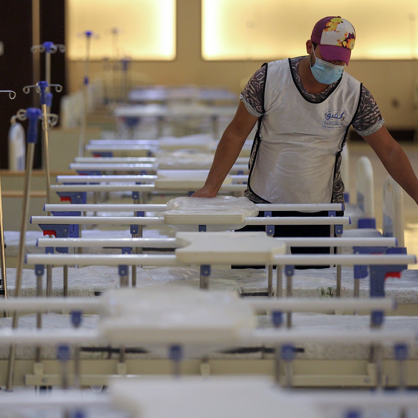 L'image montre une personne qui se trouve dans un environnement médical, probablement un hôpital ou un centre de soins. Cette personne porte un masque et un gilet, et elle est en train de s'occuper de plusieurs lits médicaux alignés. Les lits sont équipés de draps et de dispositifs intraveineux. L'atmosphère semble calme et organisée, suggérant un espace dédié aux soins des patients.