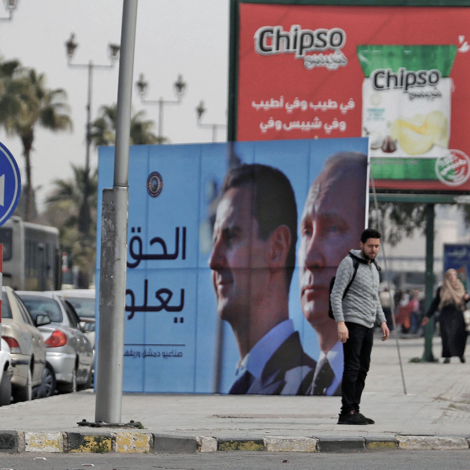 L'image montre une scène urbaine avec des panneaux de publicité. Il y a un panneau affichant des portraits de deux hommes, probablement des dirigeants, avec un message en arabe. À gauche, plusieurs personnes marchent le long de la route, tandis qu'à droite, des voitures sont garées. On peut également voir un rond-point typique et des palmiers en arrière-plan, ce qui donne un aspect typique d'une ville dans une région chaude. La publicité pour une marque de chips est visible au-dessus.