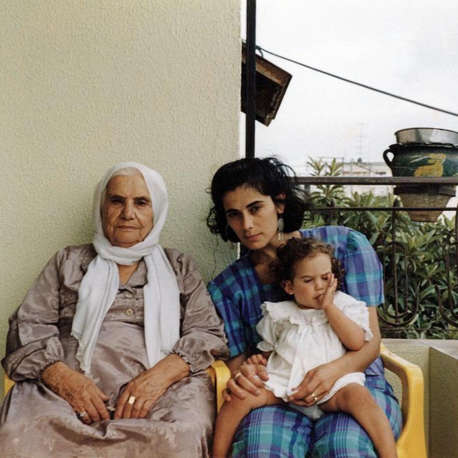 L'image montre trois femmes assises sur un balcon. À gauche, une femme âgée porte une robe grise et un foulard blanc. Au centre, une jeune femme, avec les cheveux noirs et bouclés, regarde vers l'objectif tout en tenant un enfant. L'enfant, assis sur les genoux de la jeune femme, semble jouer avec quelque chose. Le fond montre un environnement urbain avec des plantes et des éléments architecturaux. L'ambiance de l'image est chaleureuse et familiale.