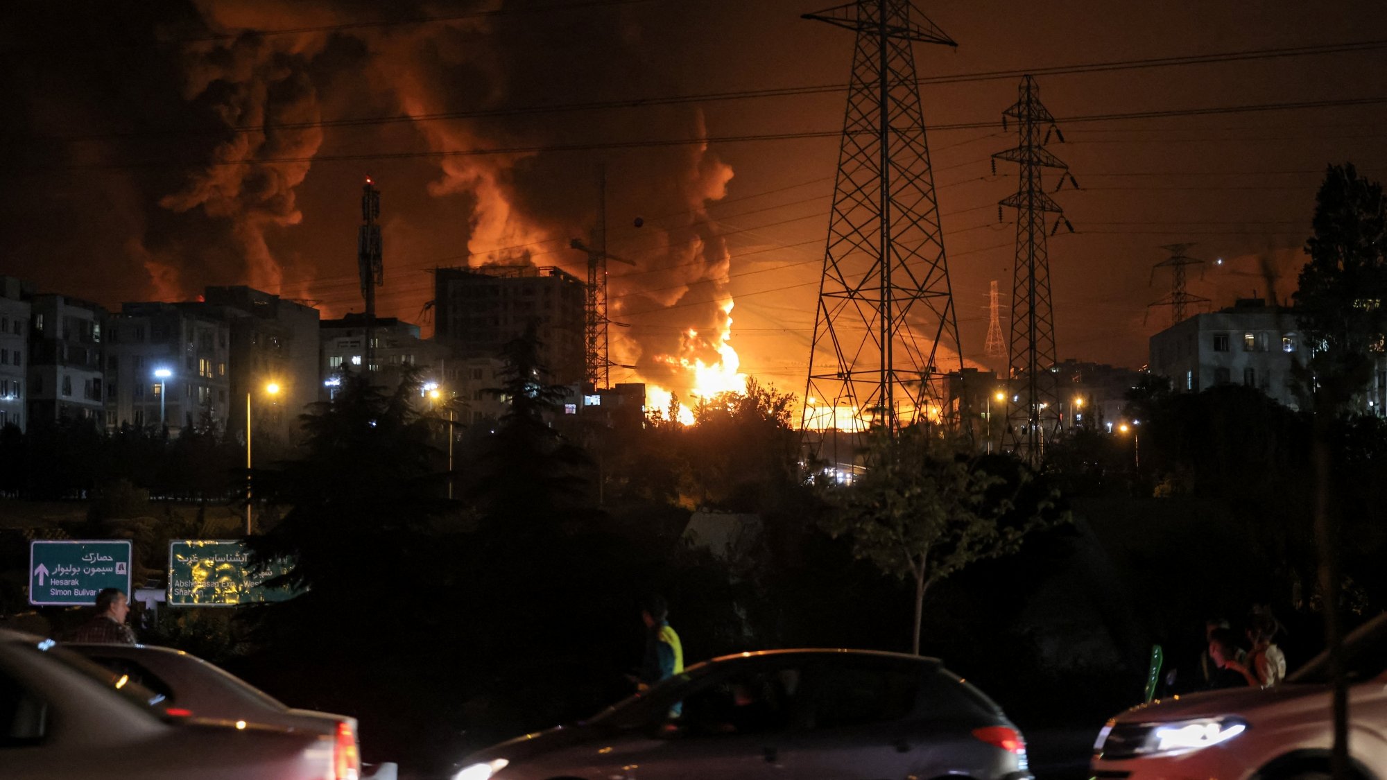 تصویری تاریک با آتش و دود بزرگ، ساختمان‌ها و تیرک‌های برق در پس‌زمینه، و خودروهای در حال حرکت.