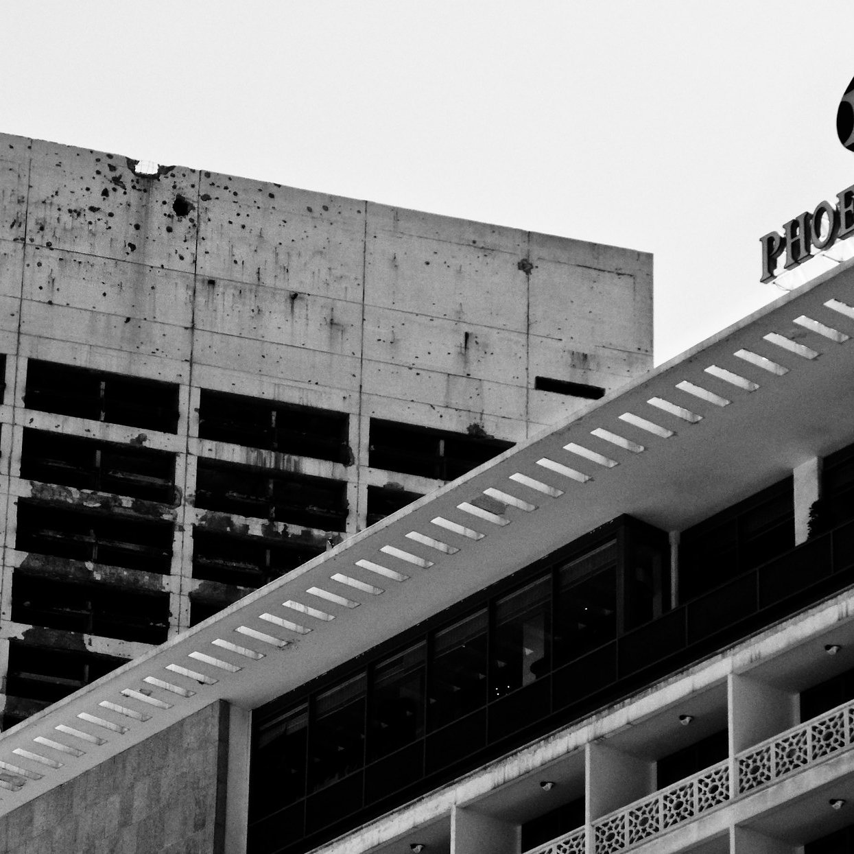 L'image montre deux bâtiments juxtaposés. À gauche, un bâtiment en béton brut et partiellement endommagé, avec des taches sombres visibles sur sa surface. À droite, un bâtiment plus moderne avec des lignes épurées et des balcons, arborant une façade blanche et des détails décoratifs. En haut à droite, le nom "Phoenix" est visible, probablement sur un panneau. L'ensemble de l'image est en noir et blanc, ce qui accentue le contraste entre les deux structures et leur état.