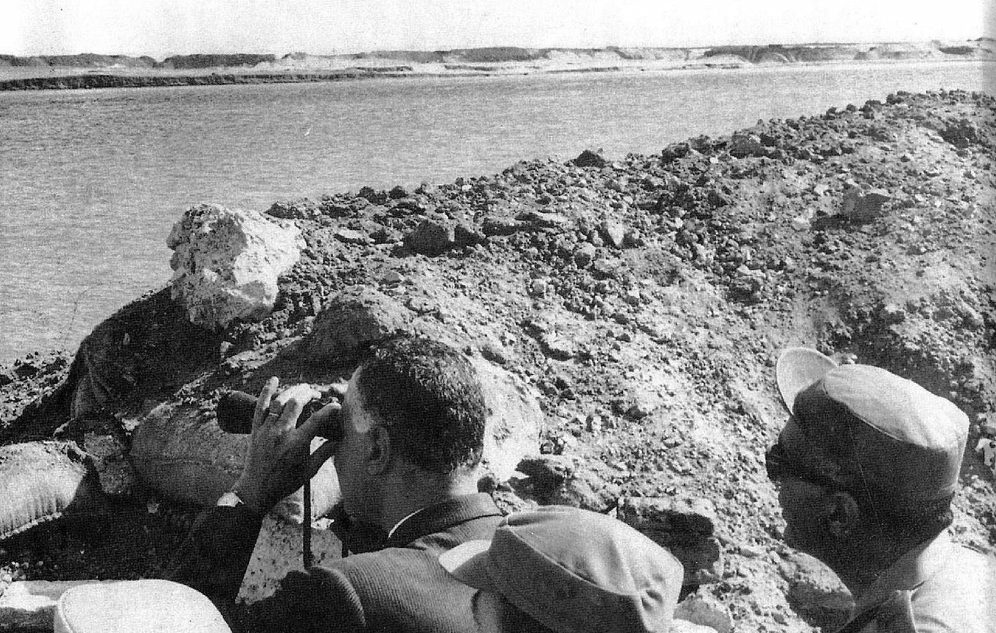 تظهر في الصورة مجموعة من الأشخاص يقفون على حافة منطقة مائية، يراقبون شيئًا ما باستخدام المنظار. يبدو أن الشخص الموجود في المقدمة مشغول بالنظر من خلال المنظار، بينما يراقبه الآخرون من حوله. الخلفية تتكون من تربة وعناصر طبيعية، مما يوحي بأن الصورة قد التُقطت في موقع استراتيجي أو أثناء زيارة ميدانية.