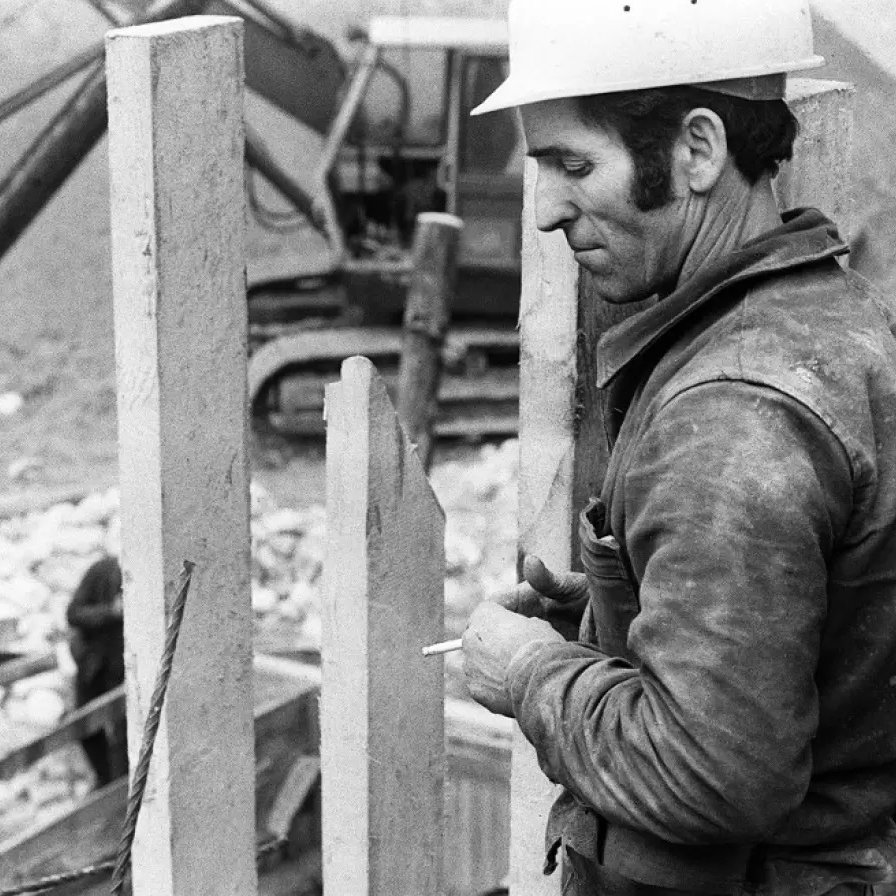 في الصورة، يظهر عامل بناء يرتدي خوذة بيضاء وهو يقف بالقرب من مجموعة من الأعمدة الخرسانية. يبدو أنه مركز على عمله، حيث يمسك بيده سيجارة. في الخلفية، يوجد عمال آخرون وأحدهم يعمل على آلة ثقيلة تُستخدم في البناء. الجو العام للصورة يتسم بالجدية والتركيز، ويظهر البيئة البنائية المحيطة والتي تشمل الأتربة والمواد الإنشائية.