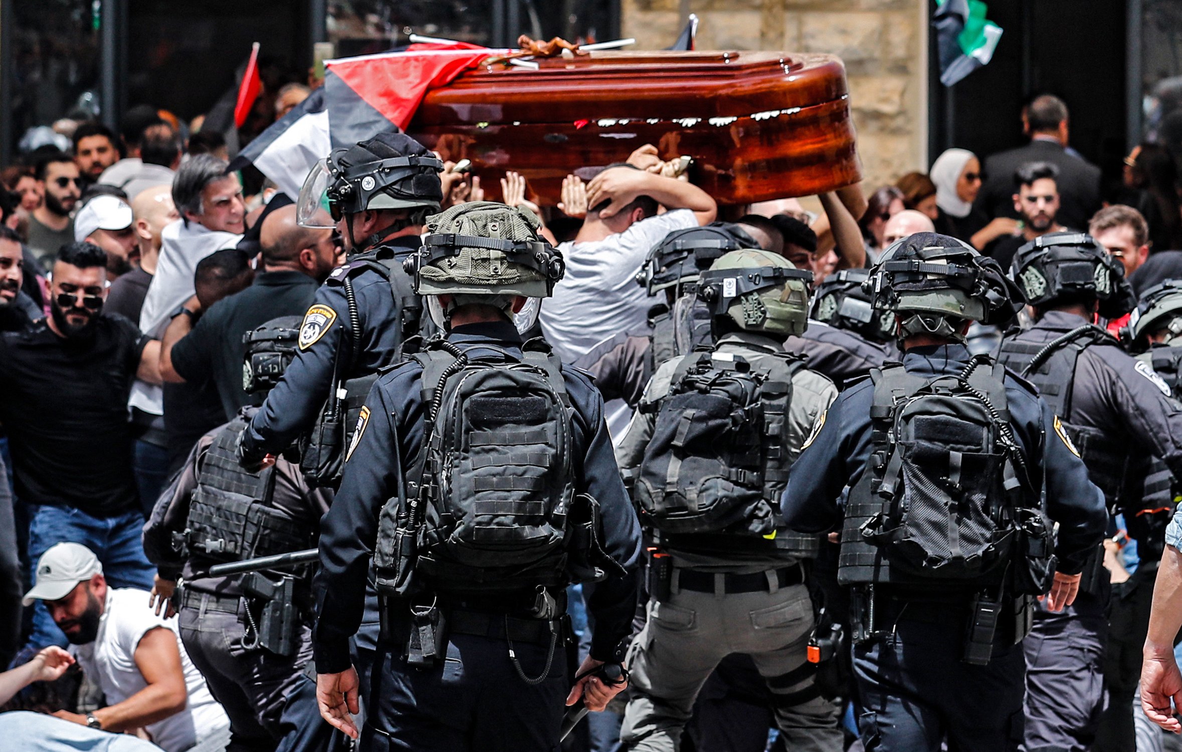 L'image montre une scène de tension où un groupe de personnes porte un cercueil en bois, tandis qu'une foule aux drapeaux palestiniens est présente. Des policiers en uniforme et équipés apparaissent en première ligne, semblant gérer la situation. L'atmosphère semble chargée d'émotion, et la scène est marquée par le contraste entre le deuil et les forces de l'ordre.