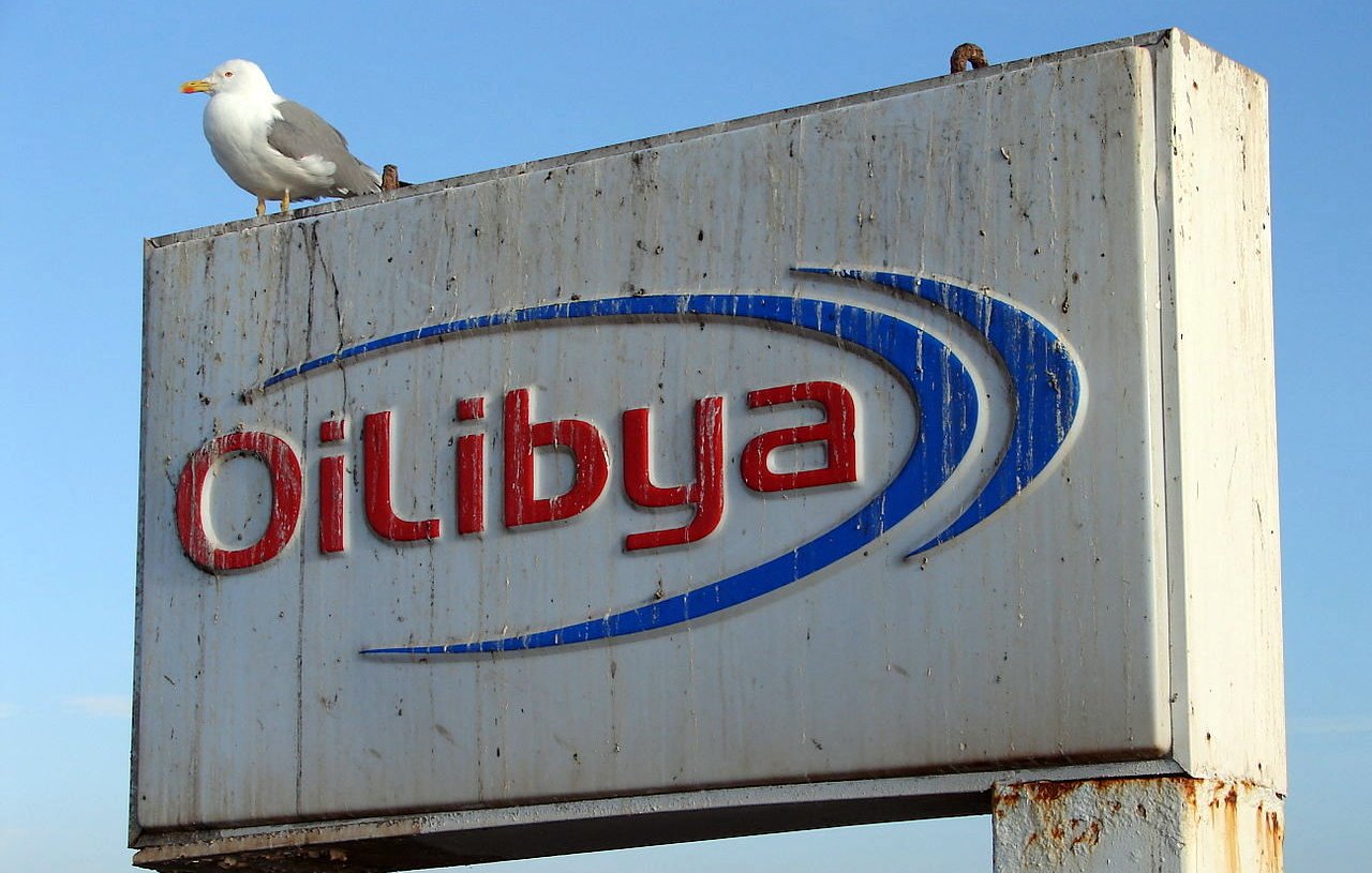 L'image montre un panneau publicitaire corrodé et usé portant le nom "Oil Libya" en lettres bleues et rouges. Au sommet du panneau, un goéland est posé. En arrière-plan, le ciel est clair, suggérant une belle journée. Le panneau semble être situé près de l'eau, probablement dans un port ou une zone côtière.