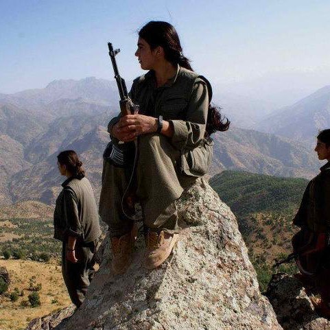 L'image montre trois personnes, probablement des combattants, dans un paysage montagneux. L'une d'elles est assise sur une pierre, tenant une arme, avec les jambes repliées. Les deux autres se tiennent debout en arrière-plan, également armées. Le décor est naturel, avec des montagnes majestueuses et une plaine en dessous, suggérant un environnement rural et sauvage. Les combattants portent des vêtements pratiques adaptés à leur environnement. L'ambiance de la scène semble sérieuse et déterminée.