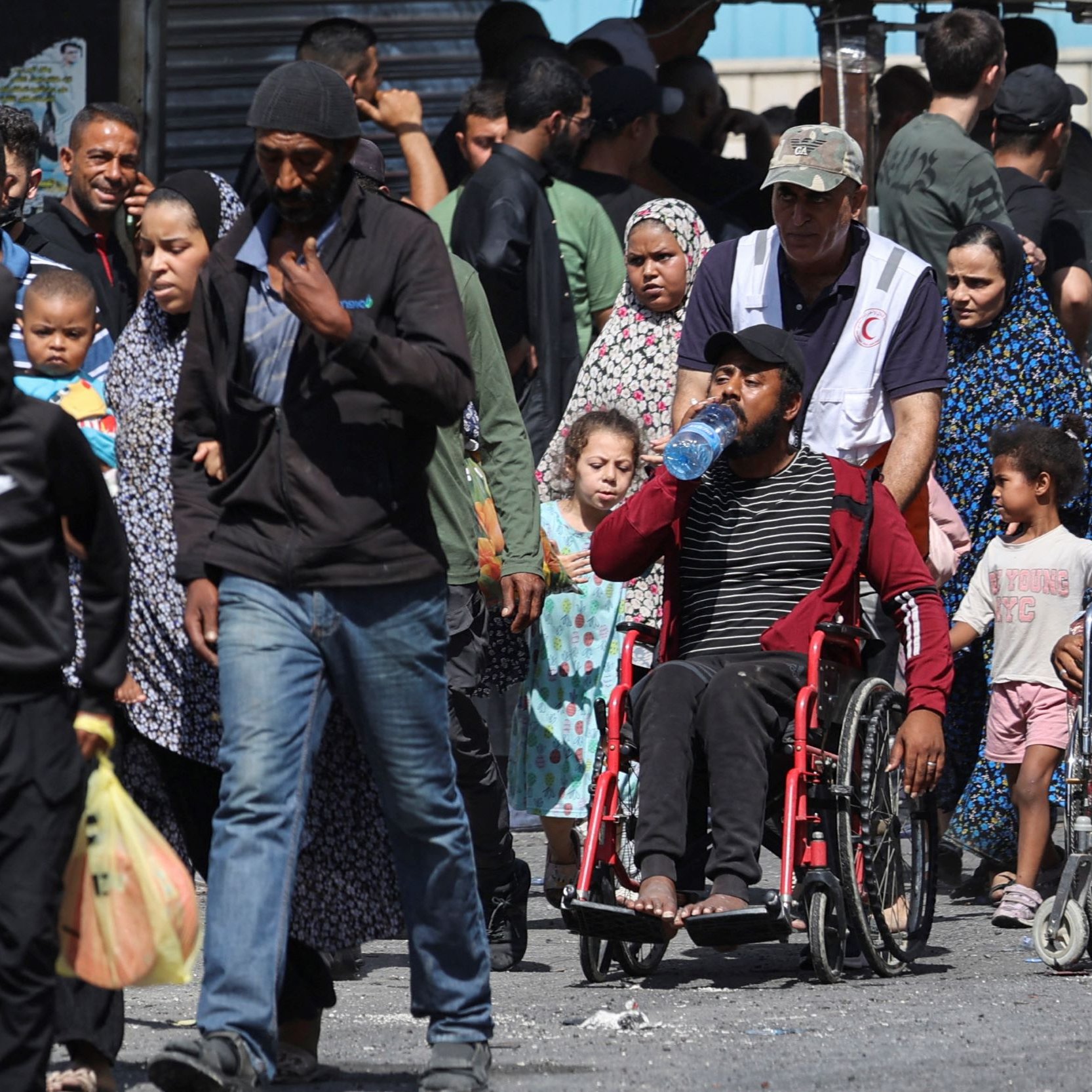 L'image montre une scène de rue animée, remplie de personnes de différents âges. On peut voir des adultes marchant, certains portant des enfants. Un homme en fauteuil roulant est également présent, buvant de l'eau. La diversité des vêtements et des expressions faciales souligne une ambiance de vie quotidienne dans un environnement urbain. En arrière-plan, on aperçoit des bâtiments et une foule d'autres personnes.
