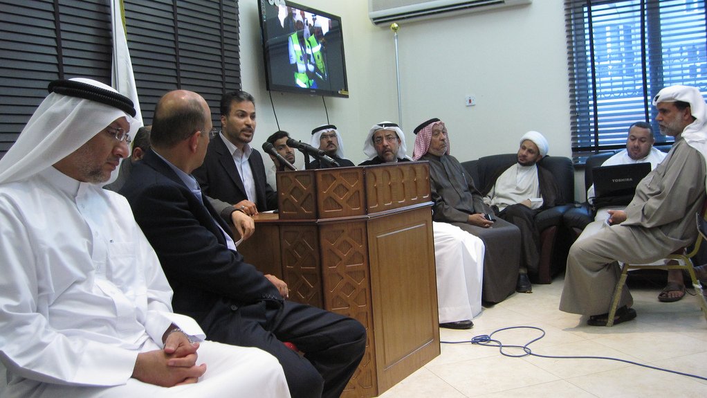 L'image montre un groupe de personnes rassemblées dans une salle. Au centre, un homme est en train de parler devant un microphone, tandis que les autres l'écoutent attentivement. Certains d'entre eux portent des vêtements traditionnels, comme des kandouras et des ghutras, indiquant peut-être un contexte culturel spécifique. À l'arrière-plan, un écran affiche probablement une présentation ou une vidéo. L'atmosphère semble sérieuse et concentrée, suggérant que la réunion pourrait porter sur un sujet important.