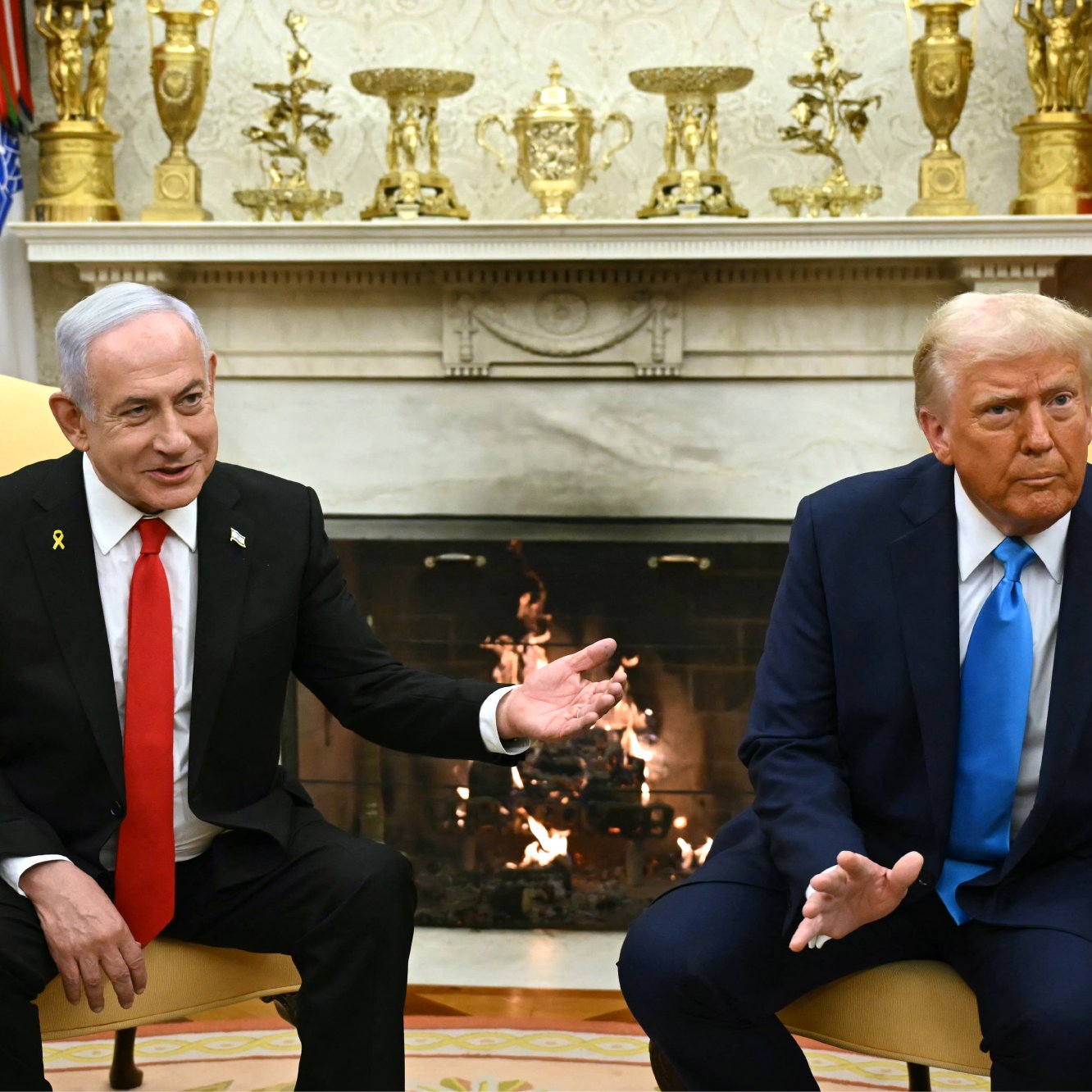 L'image montre deux hommes assis dans un bureau opulent, probablement dans le Bureau Ovale. L'un porte un costume noir avec une cravate rouge, tandis que l'autre est habillé en costume bleu avec une cravate rouge. Ils semblent engagés dans une conversation, avec des expressions sérieuses. En arrière-plan, on voit une cheminée décorée et divers objets sur des étagères. L'ambiance est formelle et politique.