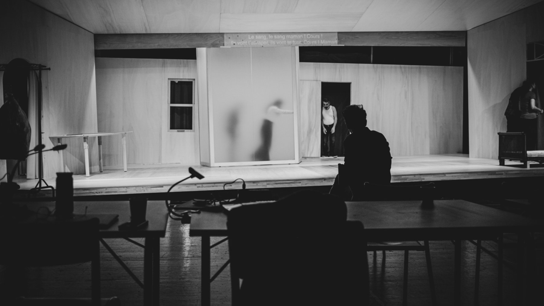 L'image montre un espace scénique avec des murs en bois clair et un éclairage tamisé. On peut apercevoir une silhouette floue à l'arrière-plan, probablement en train de se déplacer. Au premier plan, une personne est assise de dos à une table, observant la scène. L'ambiance est sombre et intimiste, suggérant un moment d'attente ou de concentration avant une représentation. Les éléments de décor, comme les fenêtres et un meuble, ajoutent à l'atmosphère théâtrale.