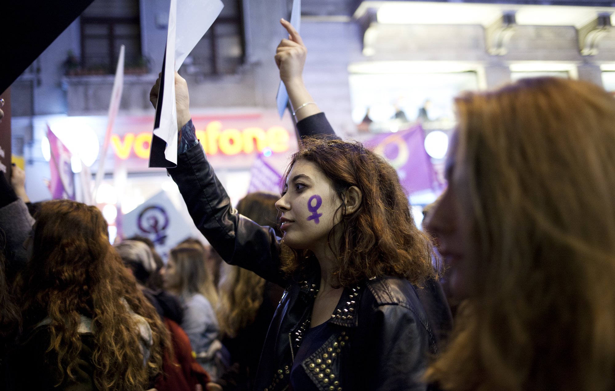 الصورة تُظهر مجموعة من المتظاهرين في تظاهرة تحتفل بحقوق المرأة. في المقدمة توجد امرأة شابة تحمل ورقة وتُظهر رمز أنثوي مرسوم على وجهها. ترتدي سترة جلدية وتبدو مفعمة بالحماس. في الخلفية، يمكن رؤية أعلام ومستندات تحمل شعارات تخص حركة النساء، مع أجواء من الحماس والتضامن بين المشاركين.