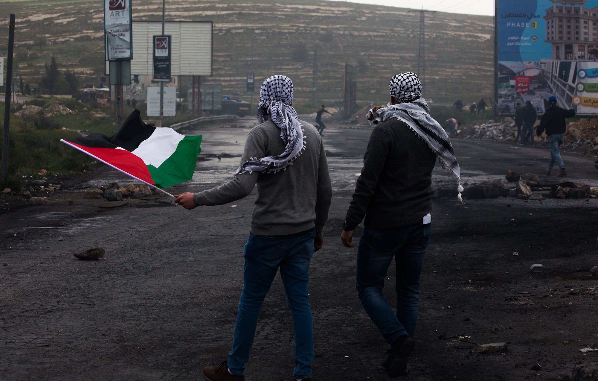 L'image montre deux hommes marchant sur une route, chacun portant un keffieh. L'un d'eux tient un drapeau palestinien, qui est composé de bandes noire, blanche, verte et rouge. L'arrière-plan présente des panneaux publicitaires, des débris et une ambiance brumeuse. Les hommes semblent se diriger vers une direction, symbolisant probablement une manifestation ou une action de revendication.