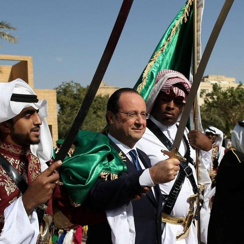 تظهر في الصورة مجموعة من الأشخاص يرتدون أزياء تقليدية. في المنتصف، يقف رجل يرتدي بدلة رسمية مع ابتسامة، محاطًا بشخصين آخرين يحمل كل منهما سيفًا. يزخر المشهد بالألوان، حيث يرتدي الشخصان الآخران ألبسة تقليدية بألوان بارزة، مع بعض الزينة الثقافية. الخلفية تضم أشجار وبعض المباني، وتبدو السماء صافية.