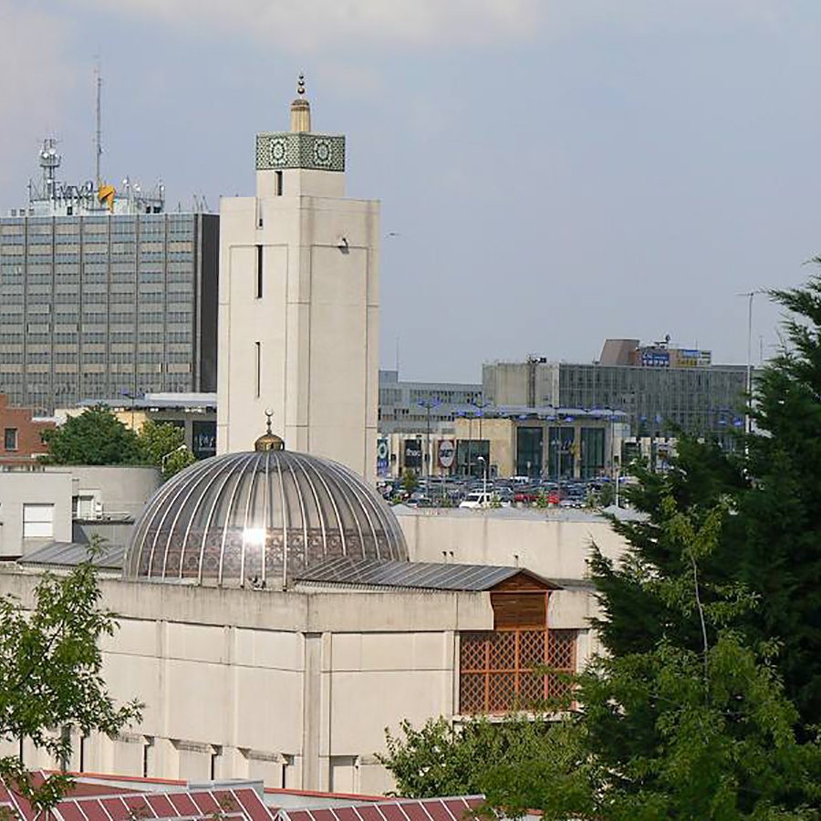 L'image montre un bâtiment architectural avec un dôme et une tour horloge, entouré d'arbres et d'autres structures urbaines en arrière-plan. On peut voir un environnement mixte, combinant des éléments d'architecture moderne et traditionnelle, avec des stations de transports et des bâtiments de bureaux. La scène est bien éclairée, suggérant une journée ensoleillée.