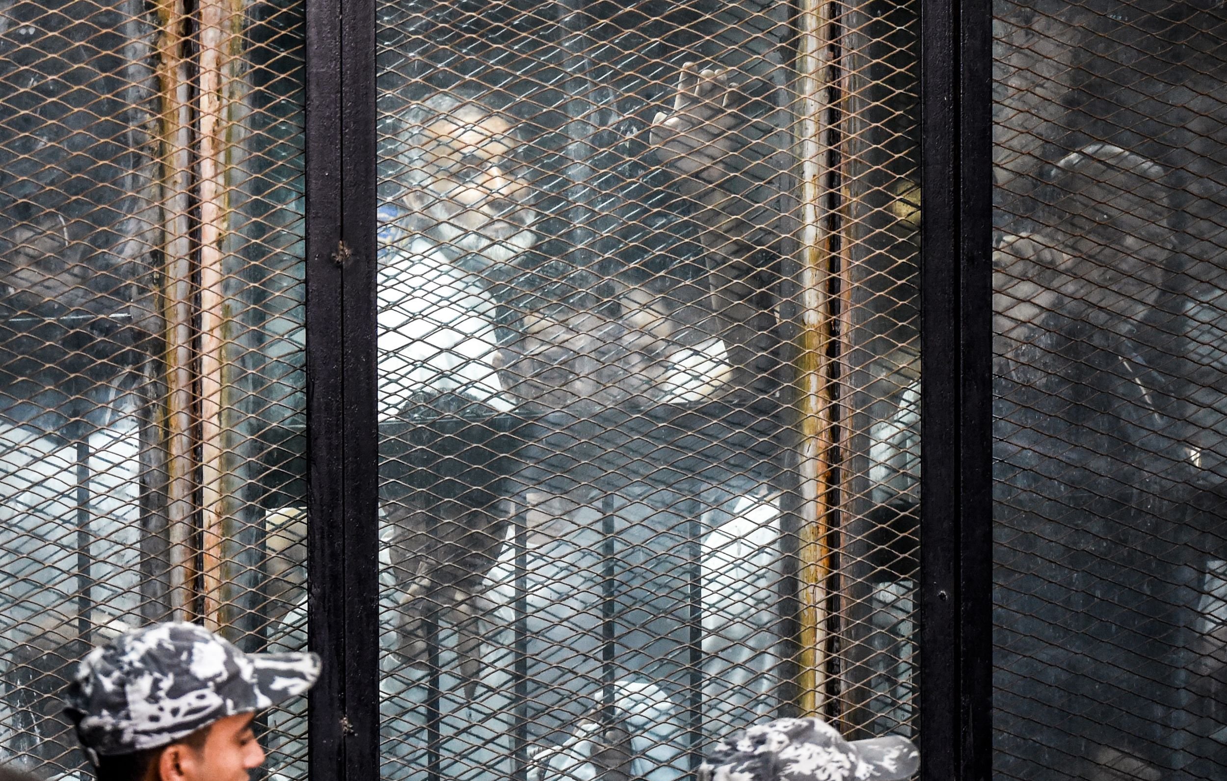 L'image montre une scène où des personnes se trouvent derrière une grille ou une clôture métallique. On aperçoit plusieurs individus, probablement en détention, qui semblent observés par des gardiens ou des forces de l'ordre en uniforme. L'atmosphère semble tendue, et il y a un mélange de visages. L'environnement donne une impression de confinement et de surveillance.