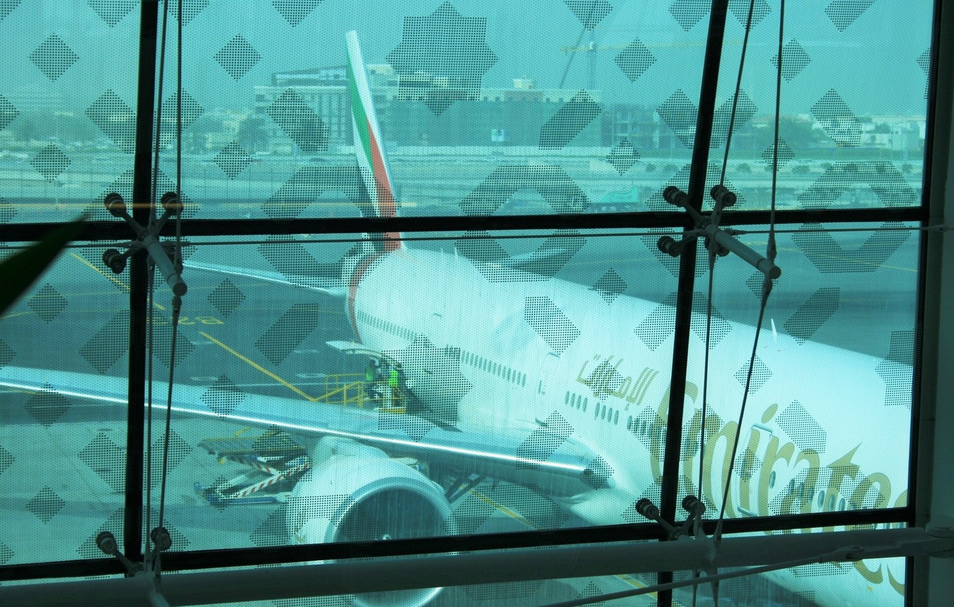 L'image montre un aéroport avec une vue à travers une grande fenêtre en verre. À l'extérieur, on peut apercevoir un avion de la compagnie Emirates, probablement un Boeing 777, stationné sur le tarmac. On distingue des travailleurs qui s'affairent autour de l'appareil, préparant son embarquement. La lumière ambiante semble assez diffuse, et il y a un léger flou qui donne une impression de profondeur. En arrière-plan, on peut voir d'autres infrastructures aéroportuaires.