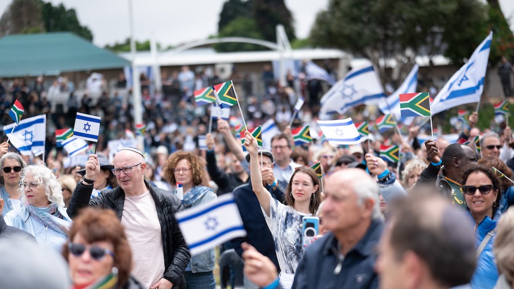 در این تصویر، گروهی از مردم در یک مراسم یا تجمع بزرگ دیده می‌شوند. آن‌ها در حال تکان دادن پرچم‌های اسرائیل و آفریقای جنوبی هستند. جمعیت شامل مردان و زنانی از تنوع سنی مختلف است و برخی از آن‌ها در حال لبخند زدن و خوشحالی دیده می‌شوند. فضای عمومی کمی ابری به نظر می‌رسد و تعداد زیادی کرسی در پس‌زمینه وجود دارد.