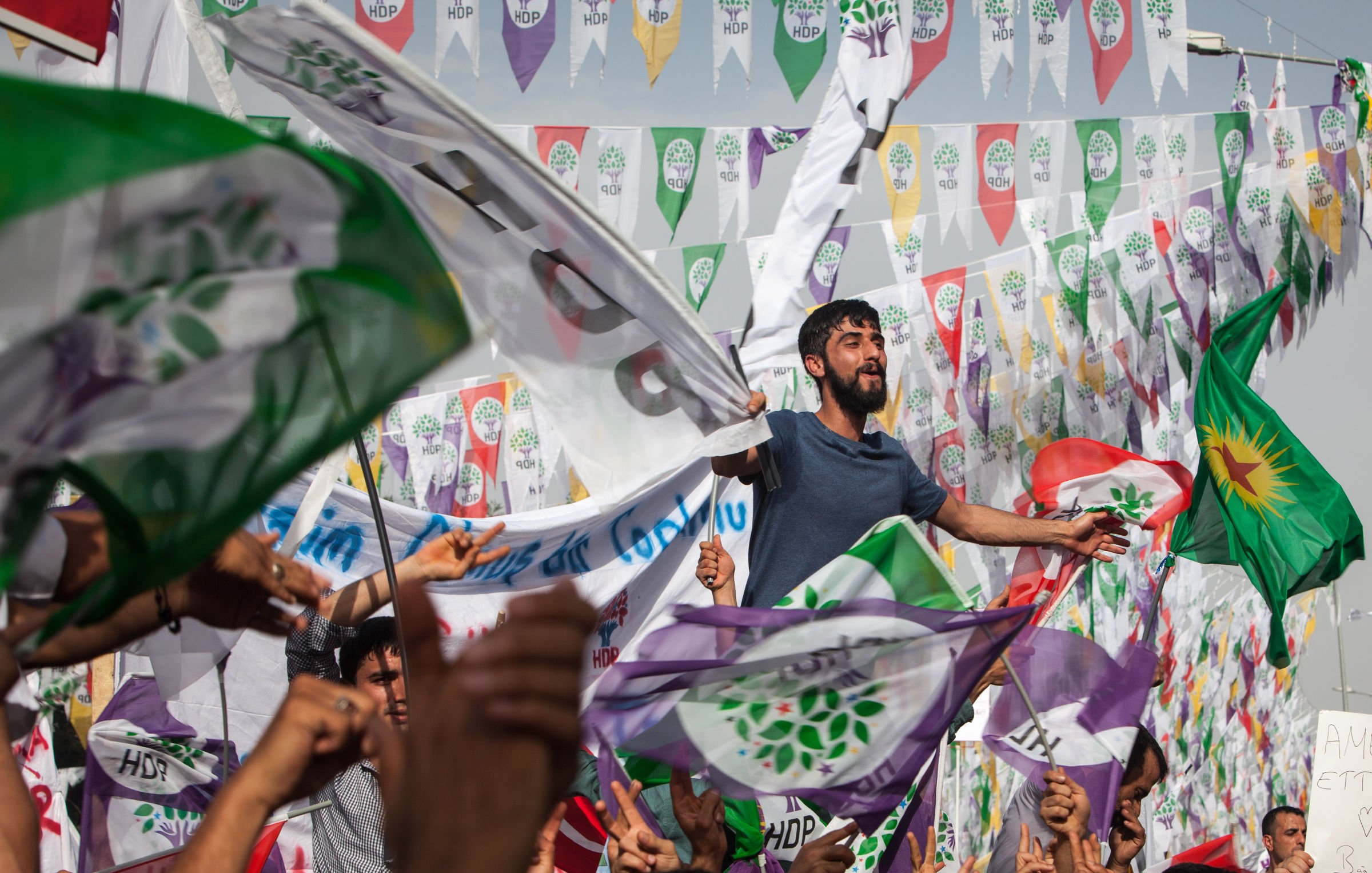 L'image montre une foule rassemblée lors d'un événement politique ou d'une manifestation. On peut voir des personnes brandissant des drapeaux, dont certains portent les couleurs et les symboles associés à un parti. L'ambiance semble festive et engagée, avec des individus visibles qui expriment leur enthousiasme, certains levant les bras ou chantant. En arrière-plan, des fanions colorés flottent, ajoutant à l'atmosphère vibrante de l'événement.