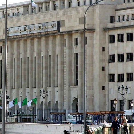 L'image montre un bâtiment majestueux avec une architecture moderne. La façade est claire, avec de grandes fenêtres et des lignes géométriques. On peut également voir des drapeaux algériens flottant devant le bâtiment. Il y a des lampadaires et une ambiance urbaine. Ce contraste entre le style architectural et le décor environnant suggère un lieu important, probablement lié aux institutions gouvernementales.