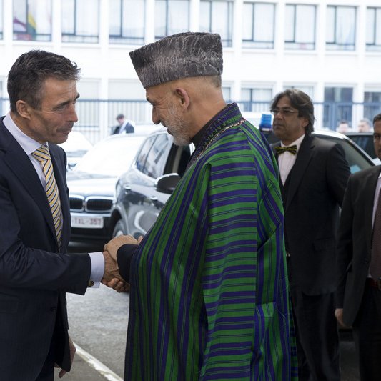 L'image montre deux hommes se serrant la main, probablement dans un cadre formel. L'un des hommes porte une tenue traditionnelle, tandis que l'autre est habillé de manière plus occidentale. À l'arrière-plan, on peut voir d'autres personnes qui semblent également habillées de façon professionnelle. L'ambiance semble être celle d'une rencontre ou d'un événement diplomatique.