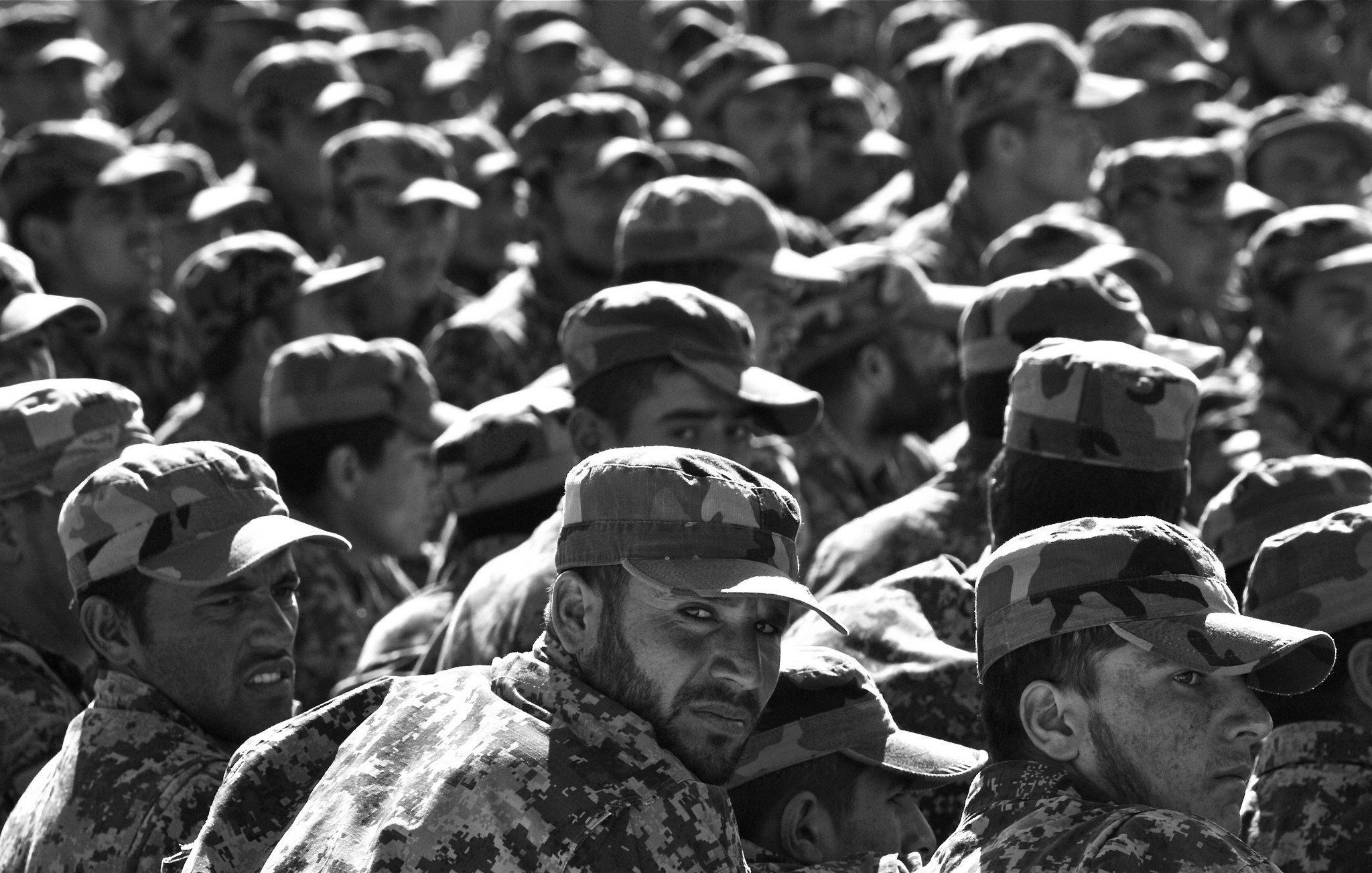 L'image montre un grand groupe de soldats en uniforme militaire, portant des casquettes assorties. Ils sont rassemblés et semblent attentifs, avec des expressions variées sur leurs visages. Le noir et blanc de la photo accentue les détails des uniformes camouflés et crée une atmosphère sérieuse, soulignant la discipline et la solidarité au sein du groupe.
