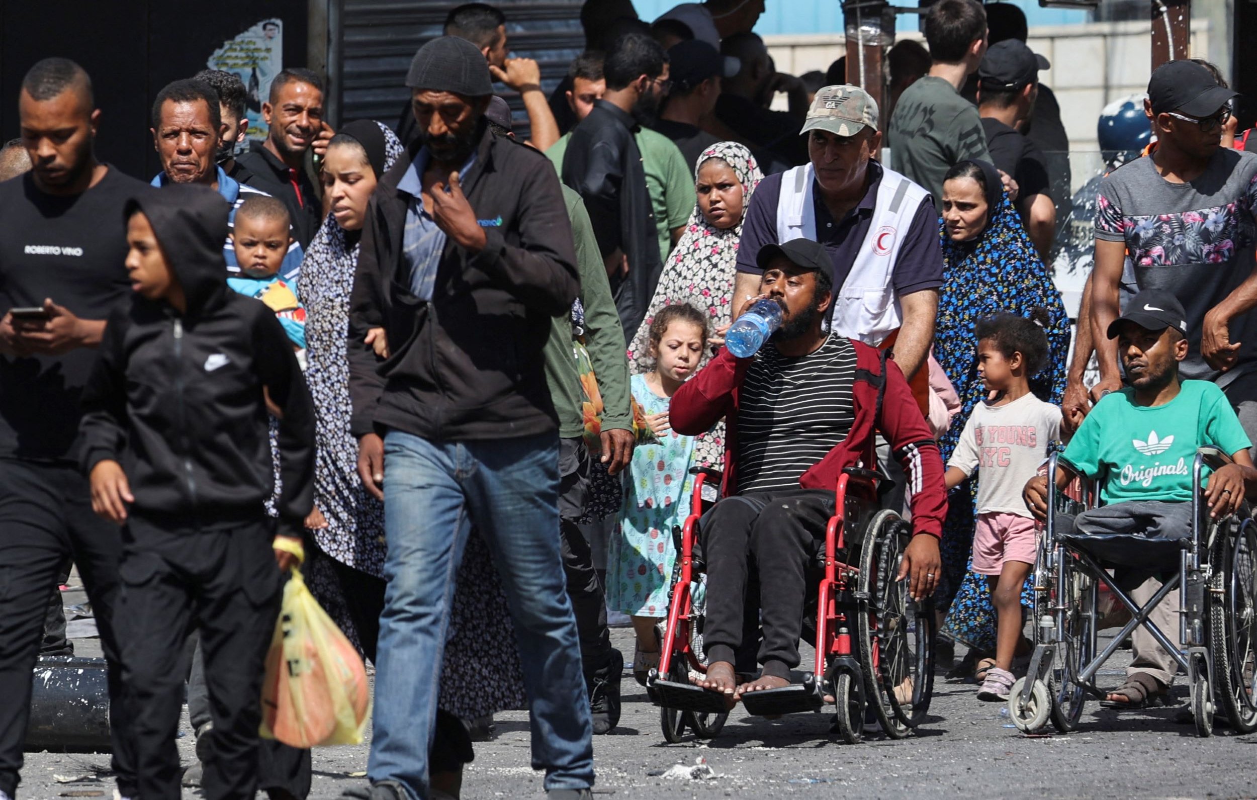 L'image montre une scène de rue animée, remplie de personnes de différents âges. On peut voir des adultes marchant, certains portant des enfants. Un homme en fauteuil roulant est également présent, buvant de l'eau. La diversité des vêtements et des expressions faciales souligne une ambiance de vie quotidienne dans un environnement urbain. En arrière-plan, on aperçoit des bâtiments et une foule d'autres personnes.