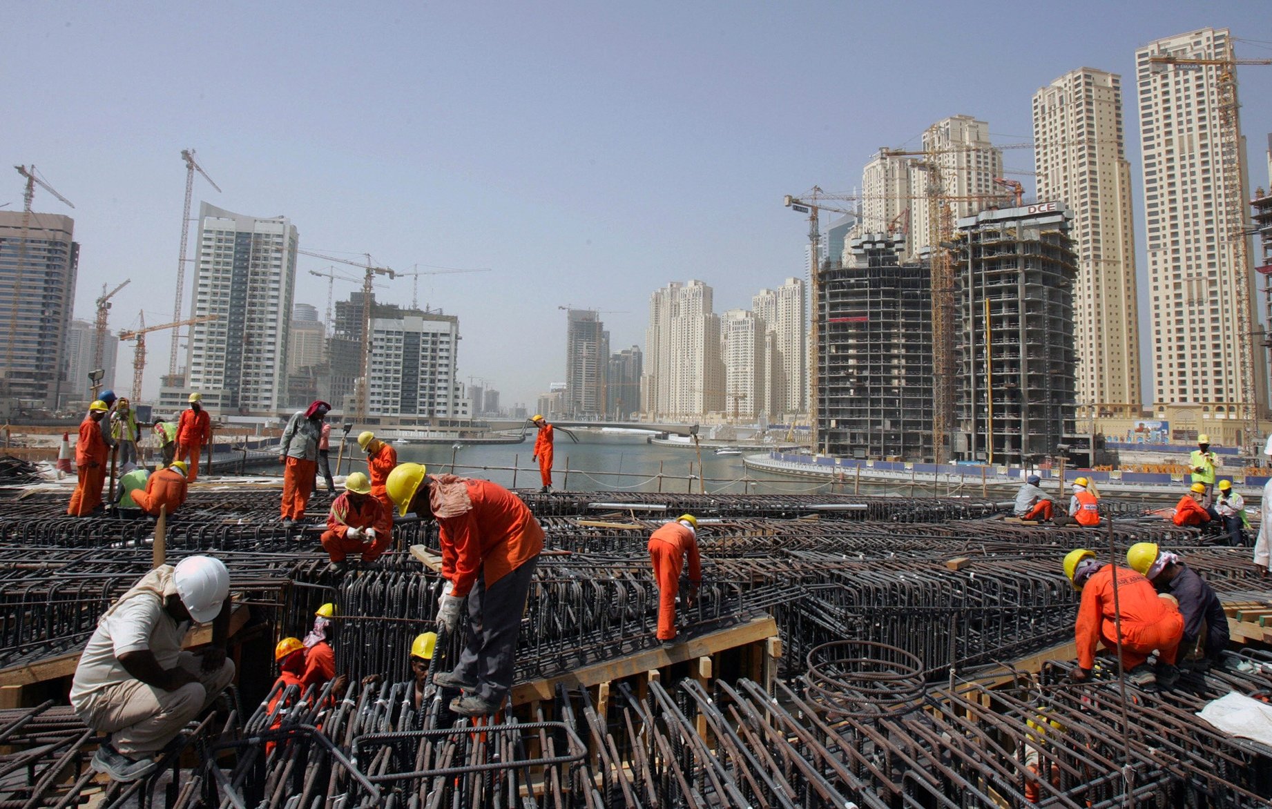 الصورة تظهر موقع بناء في منطقة حضرية حديثة. يتواجد فيها العديد من العمال يرتدون ملابس العمل البرتقالية والخوذ البيضاء. تشير الصورة إلى أن العمال مشغولون بعمليات البناء، حيث يمكن رؤية الهياكل الحديدية والرافعات في الخلفية. المباني الشاهقة الحديثة المحيطة بالموقع تدل على تطور المدينة، مع وجود بعض العمارات قيد الإنشاء. السماء تبدو صافية، ولكن هناك غبار خفيف يوحي بنشاط البناء.