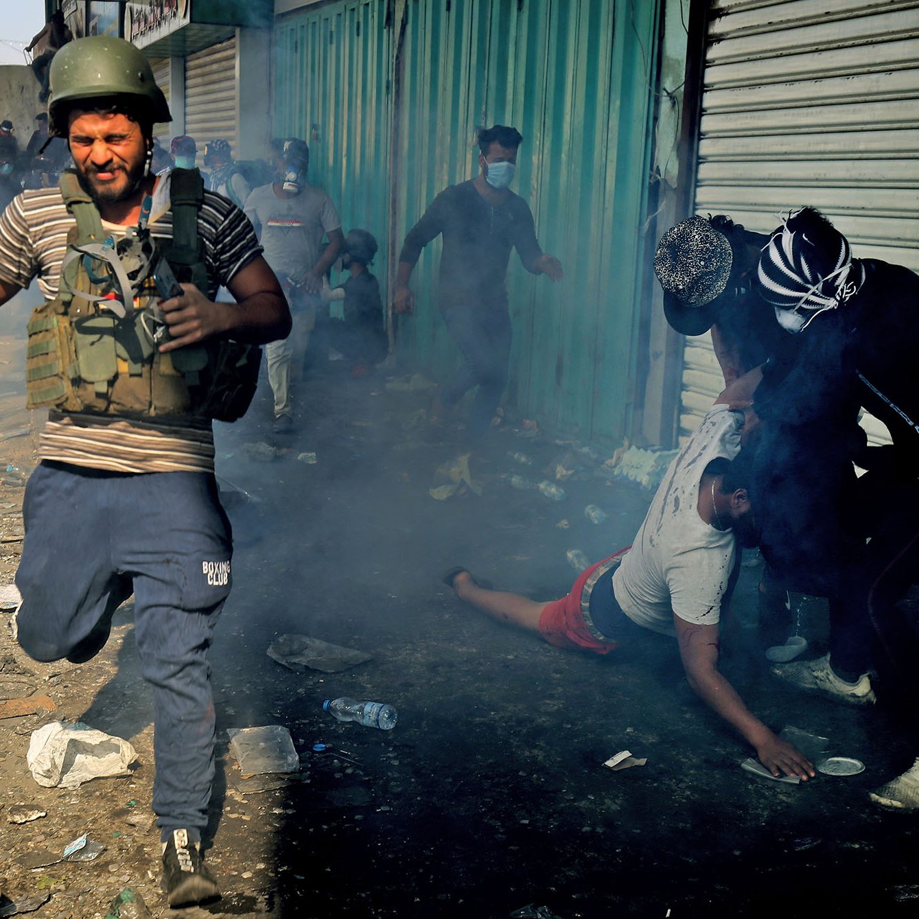 L'image montre une scène chaotique et tendue, probablement lors d'une manifestation ou d'un événement violent. Au premier plan, un homme portant un casque de protection et un gilet semble fuir rapidement, tandis qu'en arrière-plan, d'autres personnes sont engagées dans une altercation. L'atmosphère est remplie de fumée, ce qui ajoute à la tension de la situation. Le sol est jonché de débris, suggérant que l'incident a eu lieu dans un environnement urbain. Des gens semblent être en détresse, reflétant l'intensité du moment.