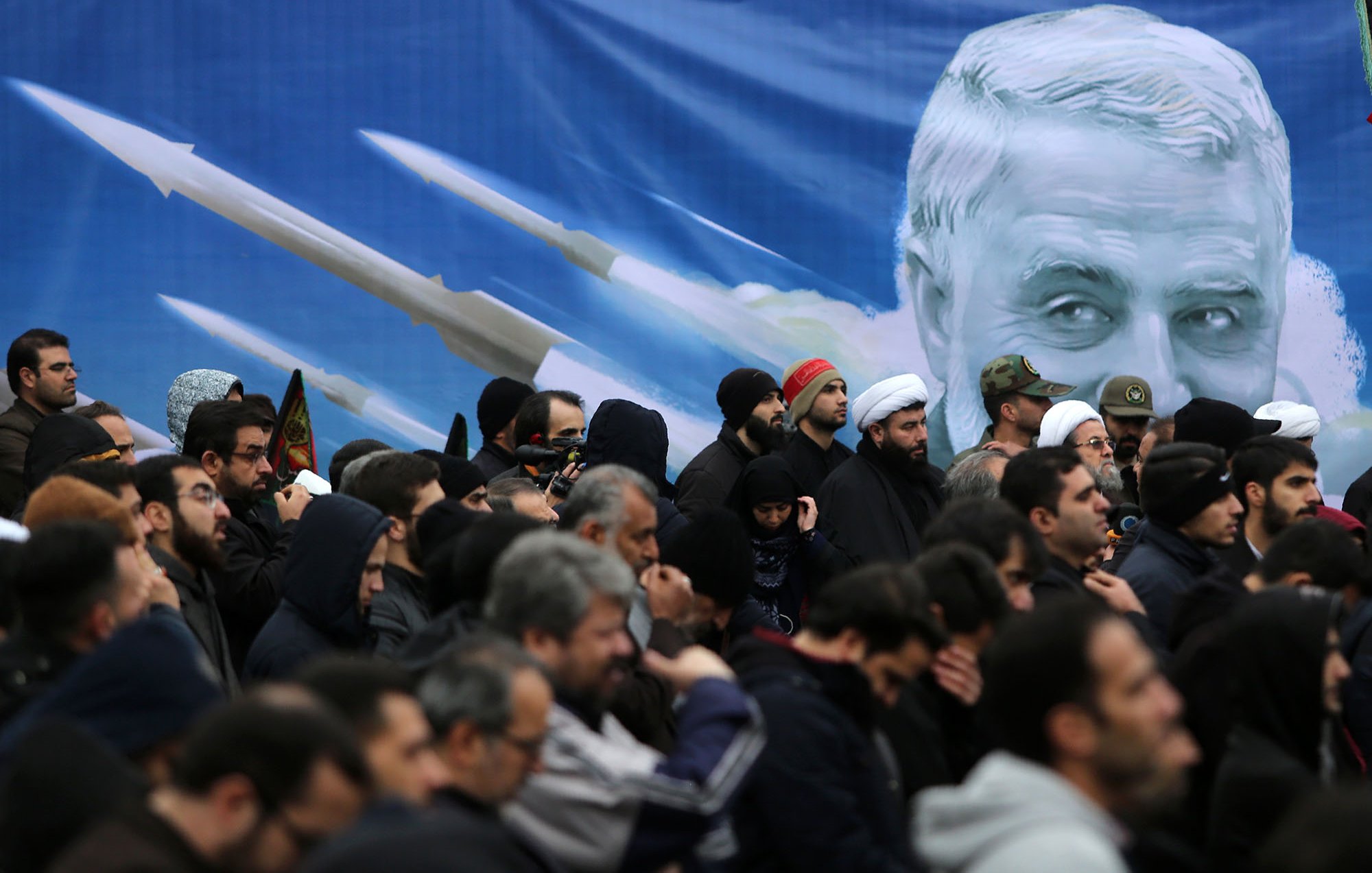 L'image montre une foule de personnes rassemblées, vraisemblablement lors d'un événement public. Les individus portent des vêtements sombres et certains semblent tenir des drapeaux ou des bannières. En arrière-plan, il y a une grande illustration d'un visage avec des images de missiles, ce qui pourrait indiquer un contexte politique ou militaire. L'ambiance générale semble sérieuse et solennelle.