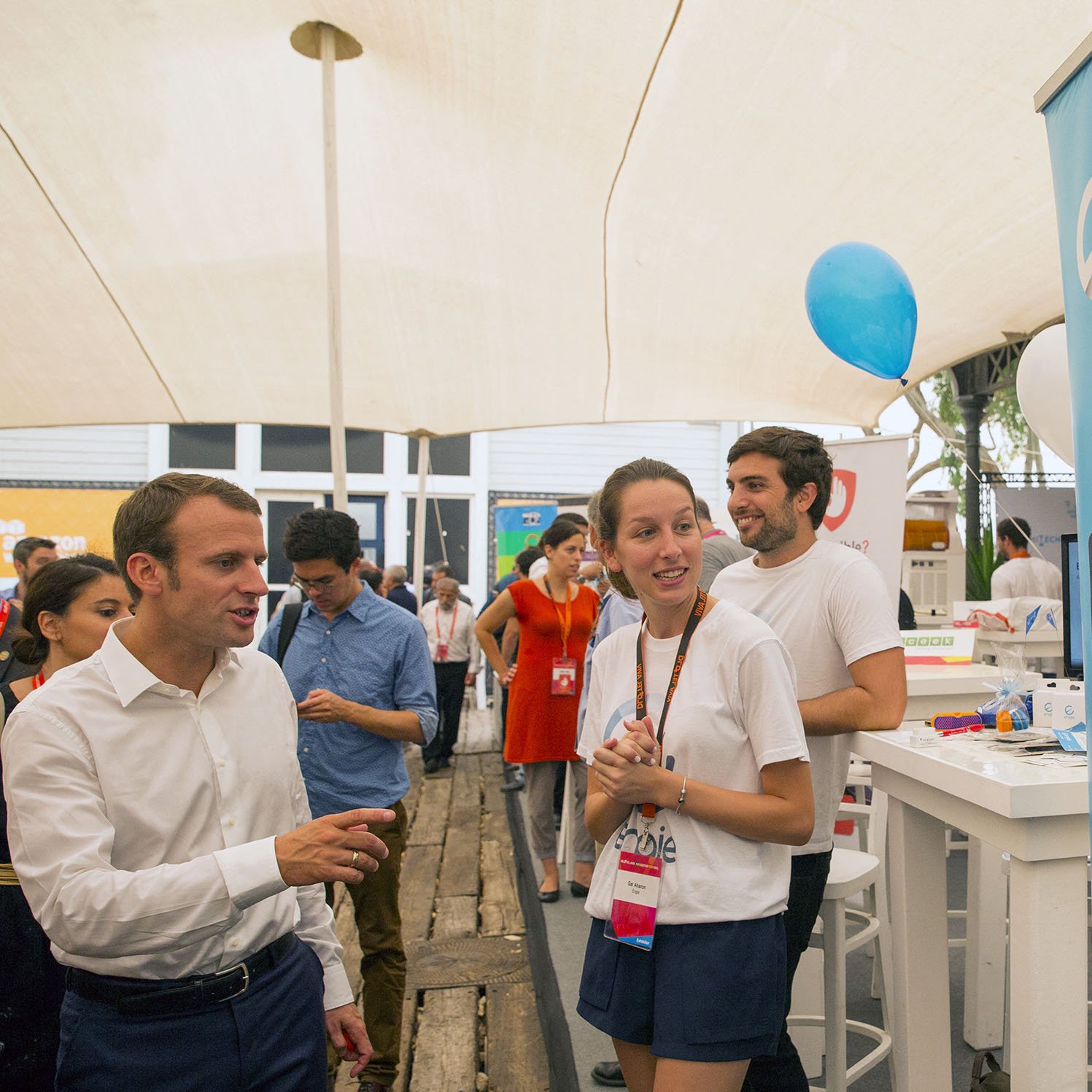 تظهر الصورة مشهداً في حدث أو معرض، حيث يتواجد عدد من الأشخاص تحت خيمة. يتمركز شخص يرتدي قميصاً أبيض، يتحدث مع شخص آخر يبدو أنه زائر. خلفهم، توجد لافتة تحمل شعار "engie" مع صور توضيحية لواجهة تطبيقات على الهواتف. يظهر في الصورة أيضاً عدد من الأشخاص الآخرين في الخلفية، وبعضهم يرتدي ملابس تحمل شارات. الجو يبدو غير رسمي ومليئاً بالنشاط.