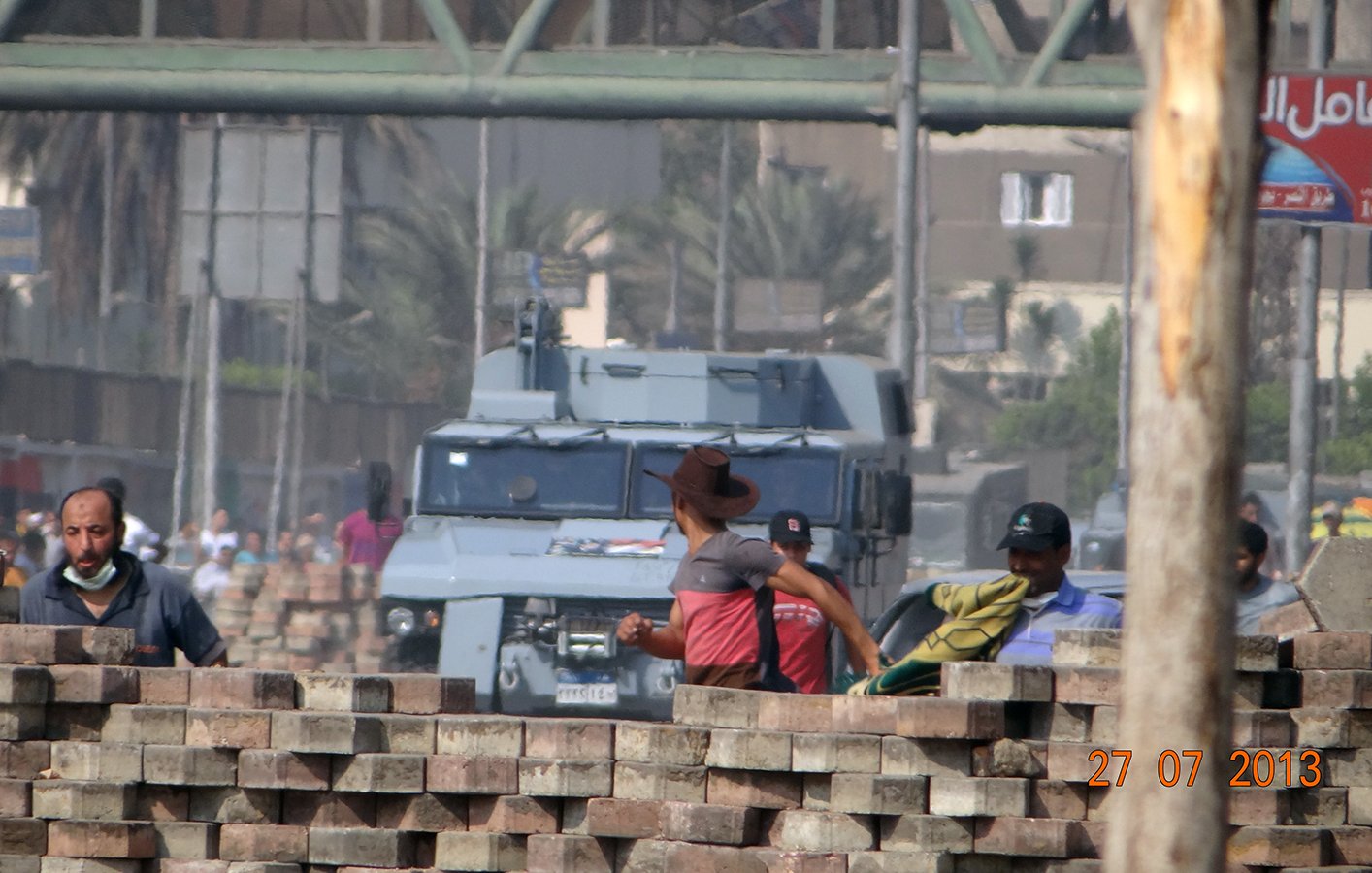L'image montre une scène urbaine probablement marquée par des tensions. Au premier plan, des hommes sont vus près d'un empilement de briques, certains portant des vêtements de travail. En arrière-plan, un véhicule blindé est visible, probablement en patrouille ou en attente. L'environnement semble indiquer une zone de conflit ou de protestation, avec des bâtiments et des plantations à proximité. La date indiquée en bas de l'image (27 juillet 2013) peut faire allusion à un événement significatif dans le contexte de la photo.