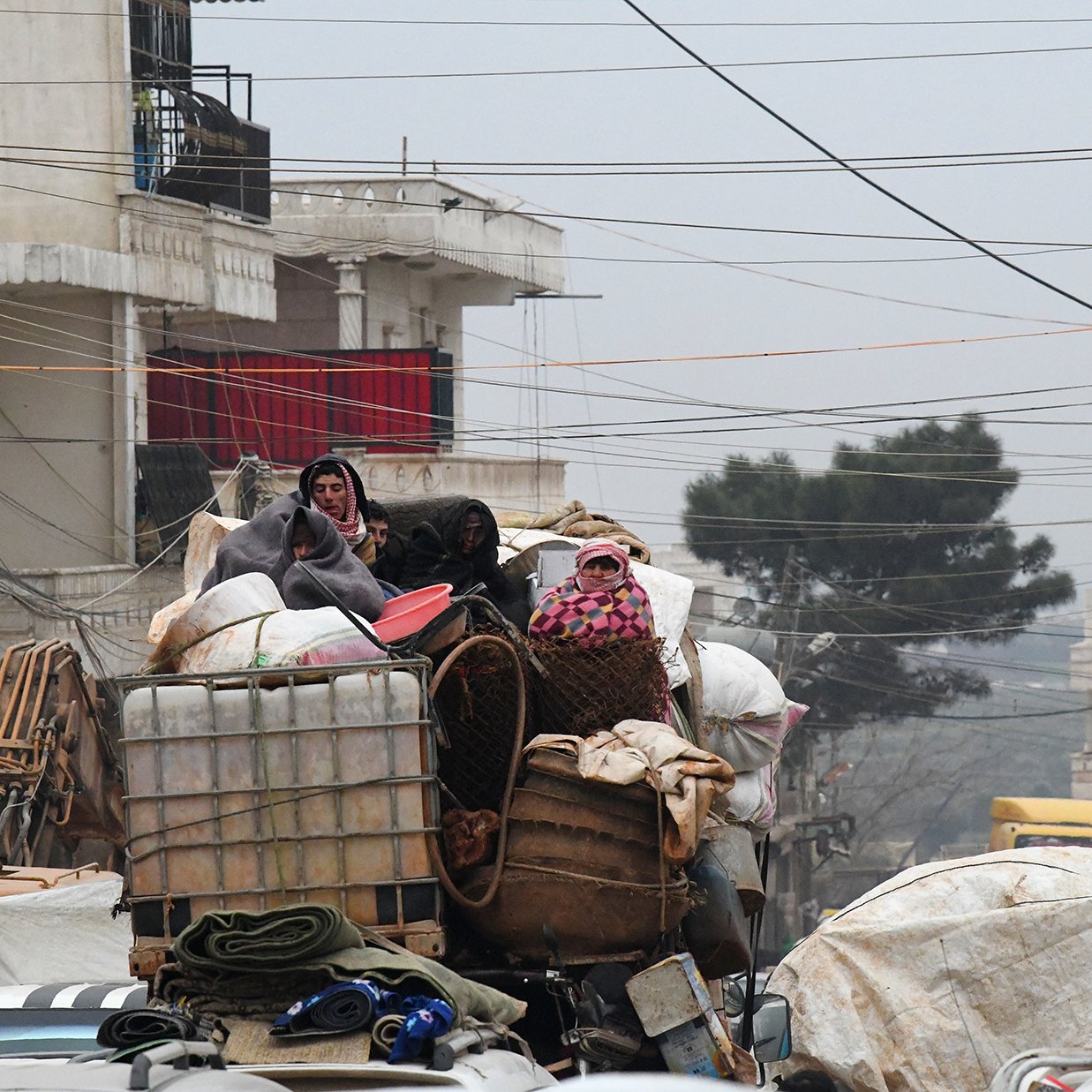 L'image montre une scène urbaine chargée, où des personnes sont perchées sur une pile de marchandises. Elles semblent se déplacer sur un véhicule surchargé, avec différents types de bagages et de matériaux. L'environnement est voilé par une lumière tamisée, peut-être due à un temps nuageux ou brumeux, et des câbles électriques traversent la scène. Les bâtiments en arrière-plan suggèrent un contexte urbain. Cette image évoque un sentiment de mouvement et de survie dans un espace urbain encombré.